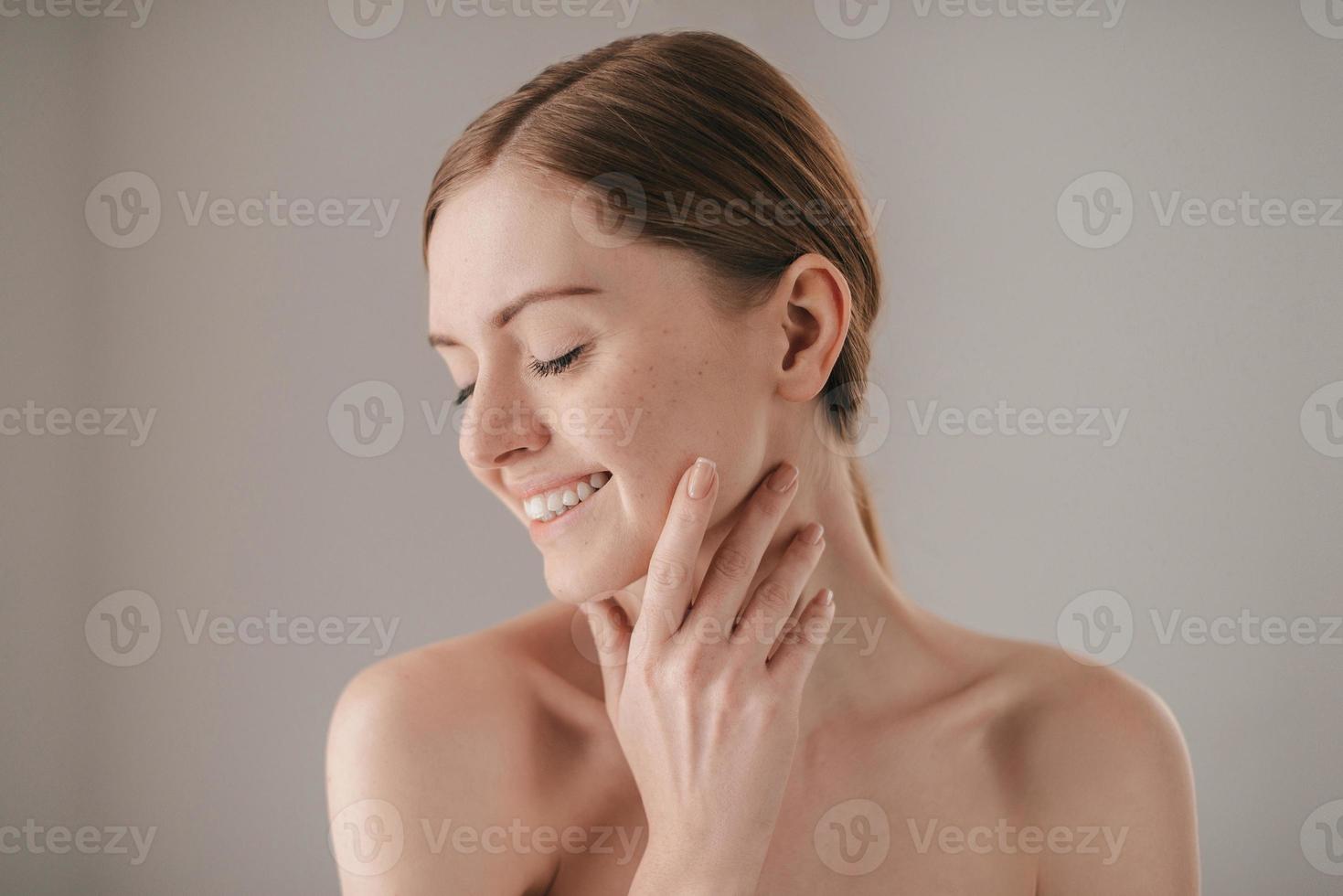 känslig vård för en lysande hud. porträtt av rödhårig kvinna med fräknar förvaring ögon stängd och leende medan rörande henne ansikte och stående mot grå bakgrund foto