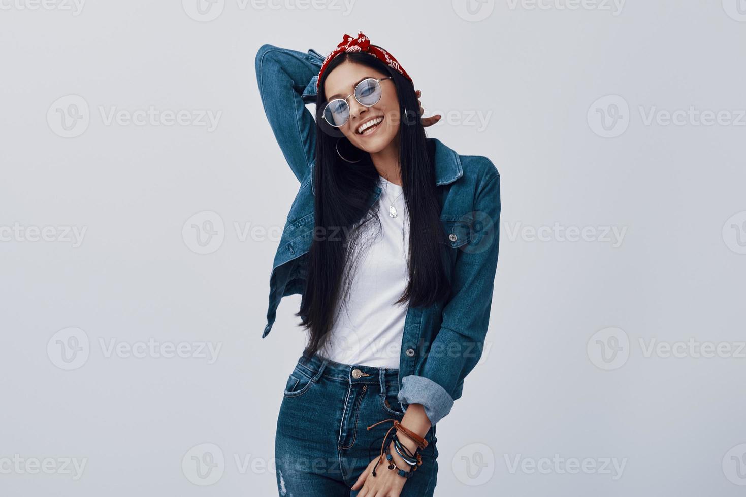 attraktiv ung kvinna i glasögon och bandana leende medan stående mot grå bakgrund foto
