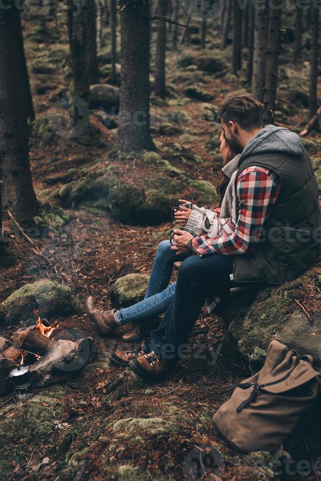 reser med älskad. skön ung par innehav koppar medan uppvärmningen upp nära de lägereld foto