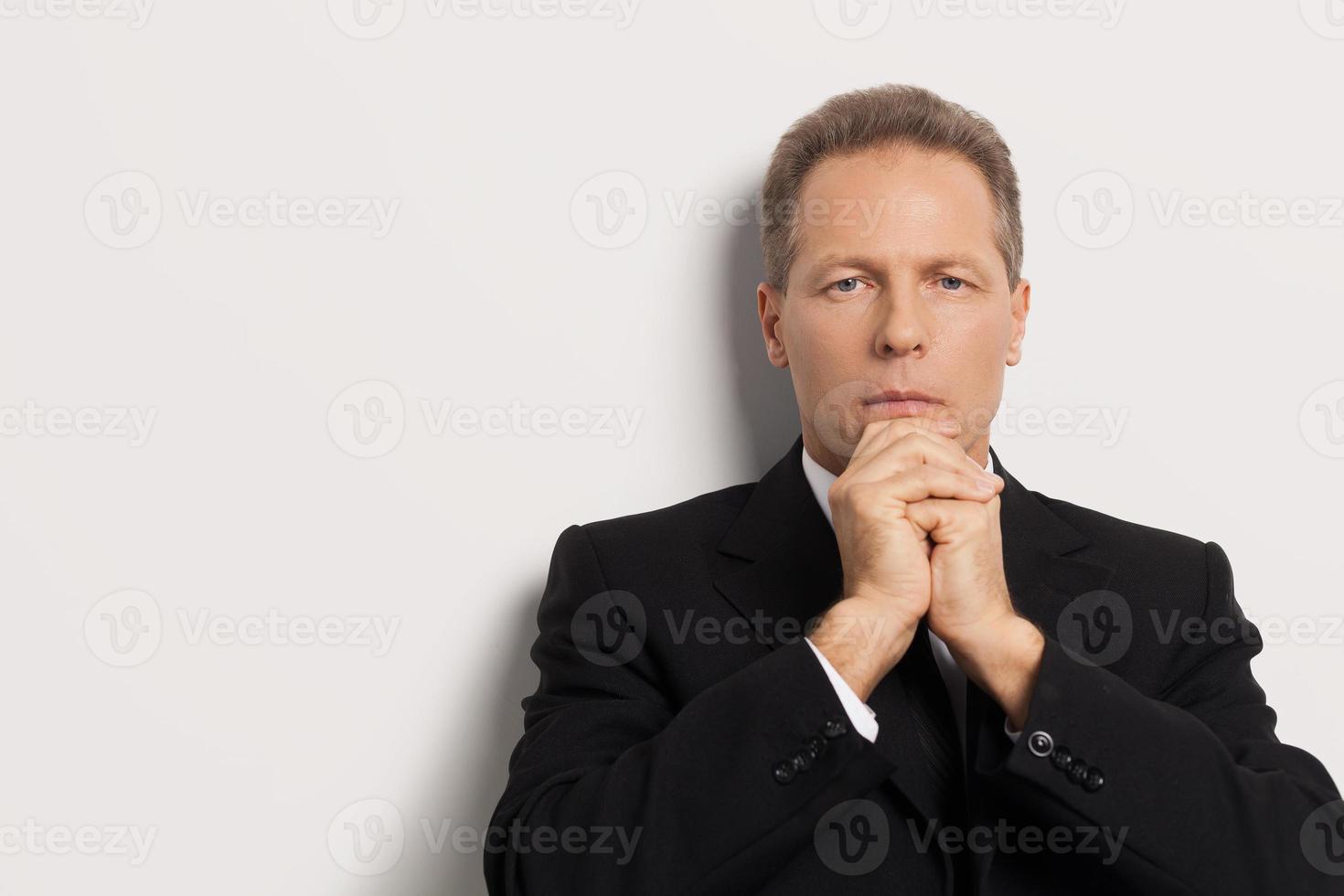 omtänksam närma sig till företag. omtänksam senior man i skjorta och slips innehav händer på haka och ser på du medan isolerat på grå bakgrund foto