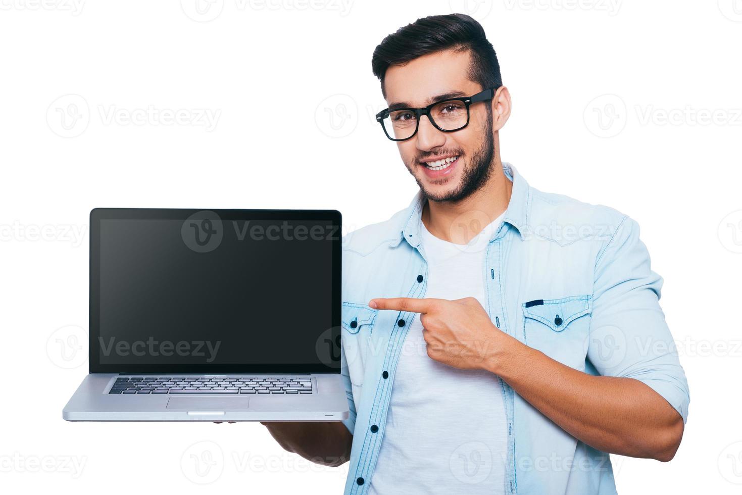 detta är Vad du behöver självsäker ung indisk man bärande bärbar dator på hand och pekande den medan stående mot vit bakgrund foto