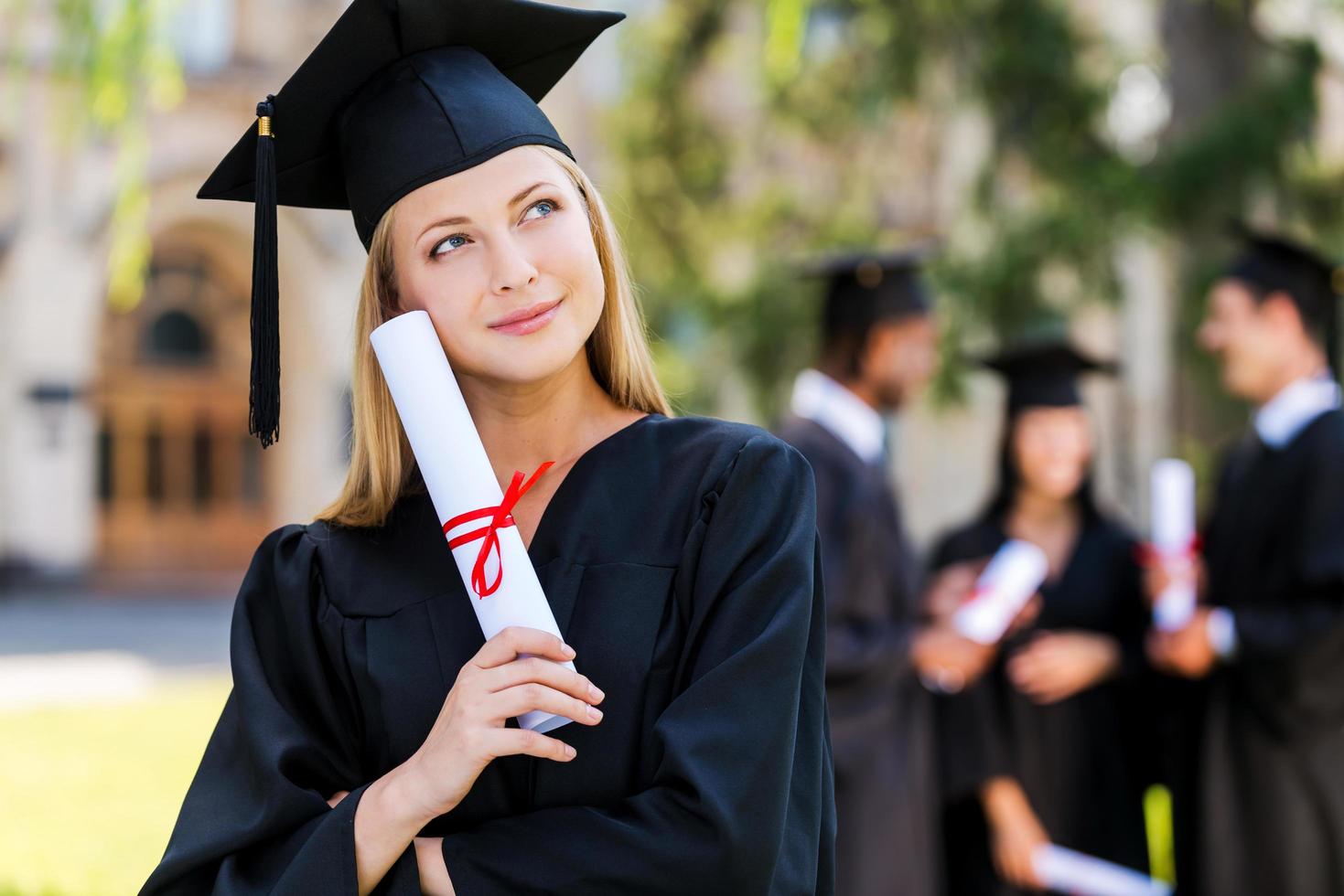 drömma av en ljus framtida. omtänksam ung kvinna i gradering kappor innehav diplom och ser bort medan henne vänner stående i de bakgrund foto