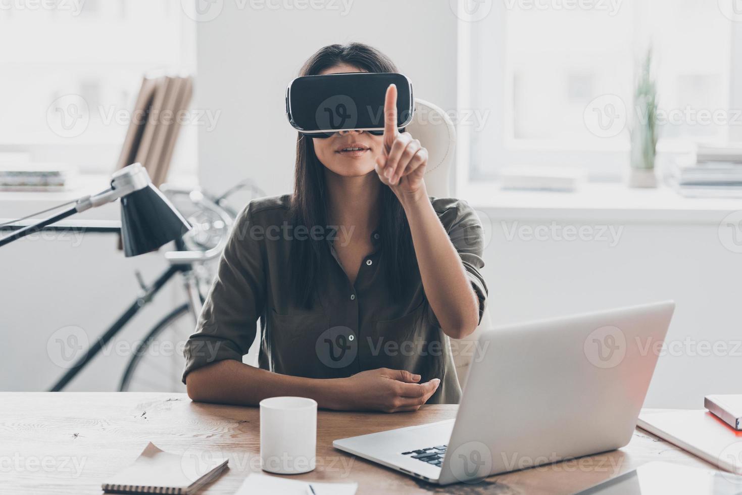 vr teknik. självsäker ung kvinna i virtuell verklighet headsetet pekande i de luft medan Sammanträde på henne arbetssätt plats i kontor foto