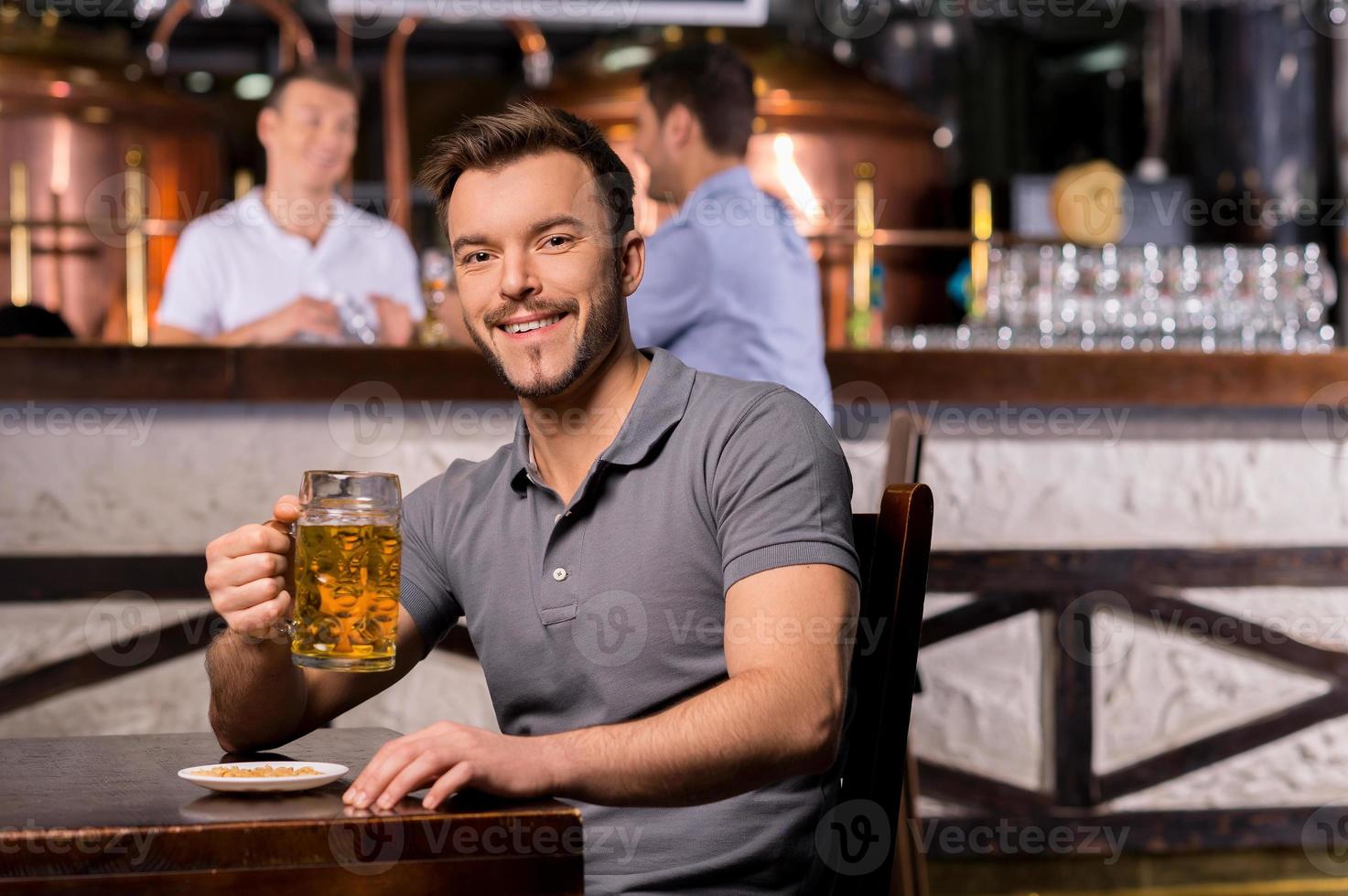 man i öl pub. glad ung man innehav en öl råna och leende medan Sammanträde i bar foto