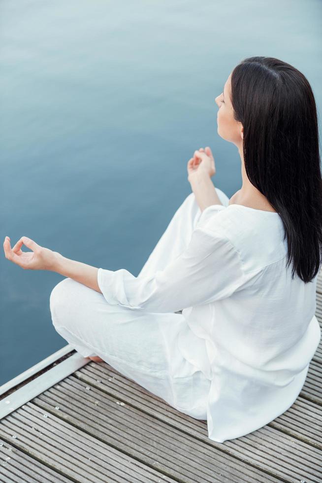 morgon- meditation. topp se av skön ung kvinna i vit Kläder mediterar medan Sammanträde på de kajkant foto