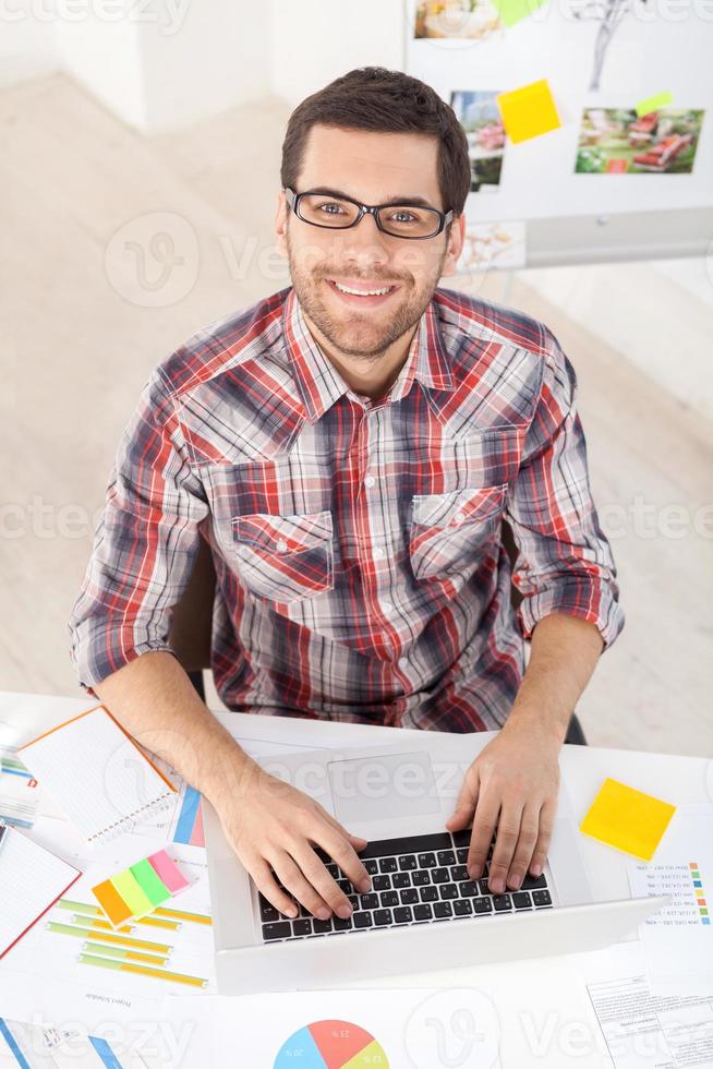 kreativ verkställande. topp se av stilig ung man i glasögon arbetssätt på dator och leende på kamera medan Sammanträde på hans arbetssätt plats foto