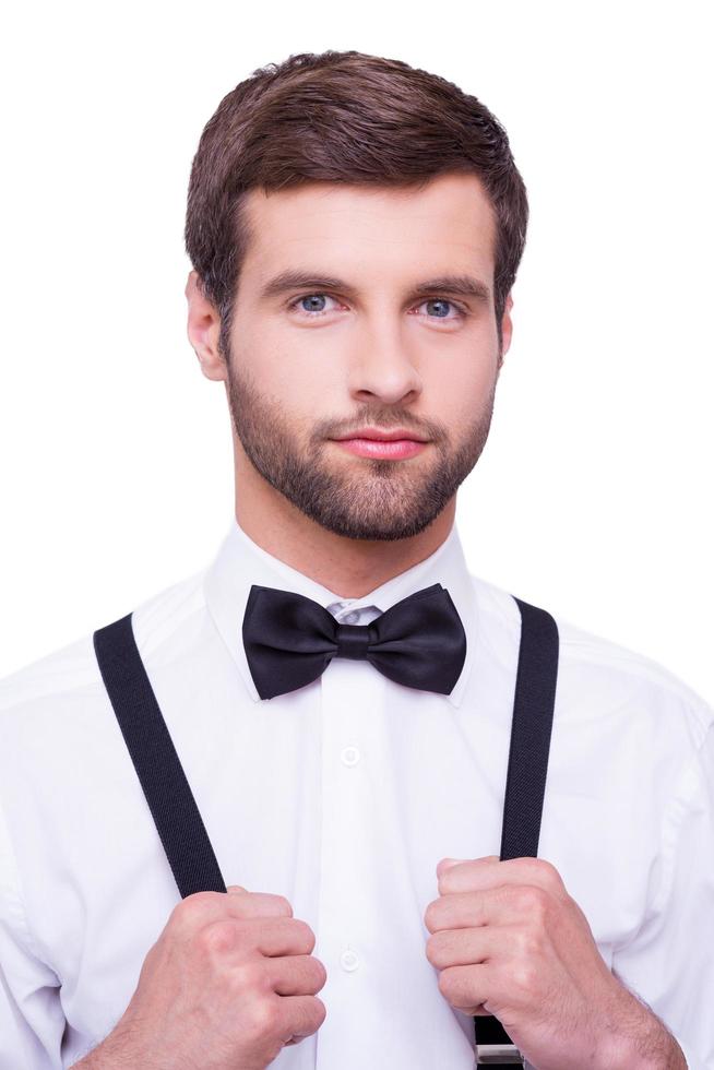 elegant och stilig. porträtt av stilig ung man i vit skjorta och rosett slips justeras hans hängslen och ser på kamera medan stående isolerat på vit foto
