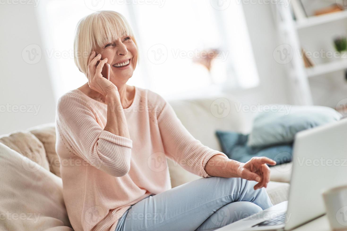 Lycklig till höra du skön senior kvinna talande på henne smart telefon och leende medan avkopplande på de soffa på Hem foto