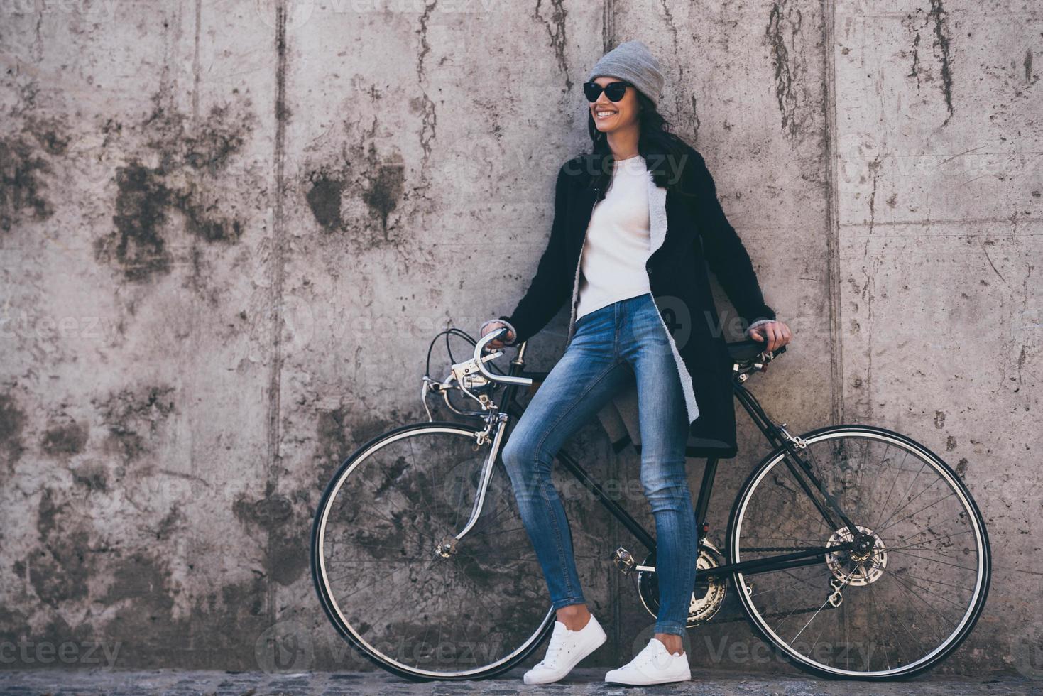 den där var bra rida skön ung kvinna i solglasögon innehav händer på henne cykel och ser bort med leende medan stående mot betong vägg utomhus foto