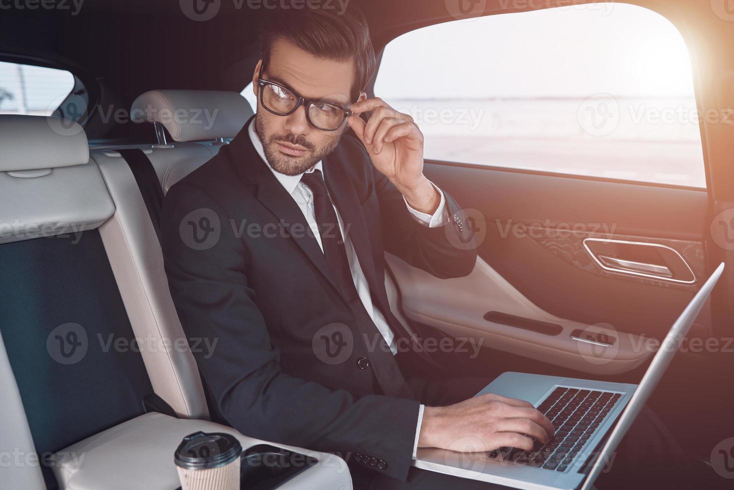 planera hans företag. omtänksam ung man i full kostym arbetssätt använder sig av bärbar dator och justeras hans glasögon medan Sammanträde i de bil foto