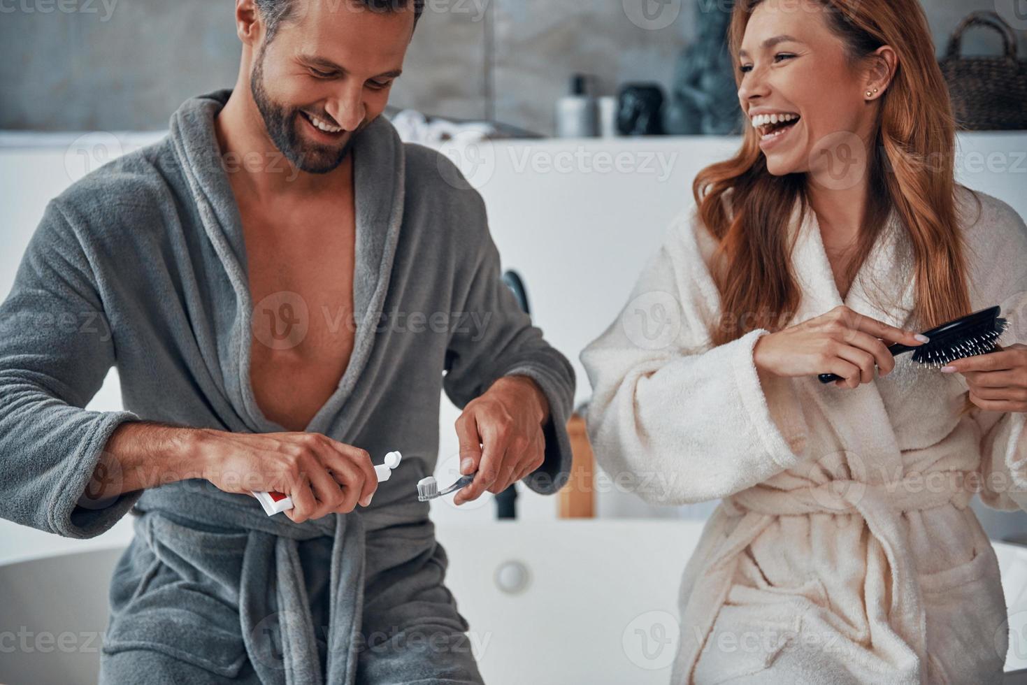 kärleksfull ung par i morgonrockar skrattande medan håller på med morgon- rutin- foto