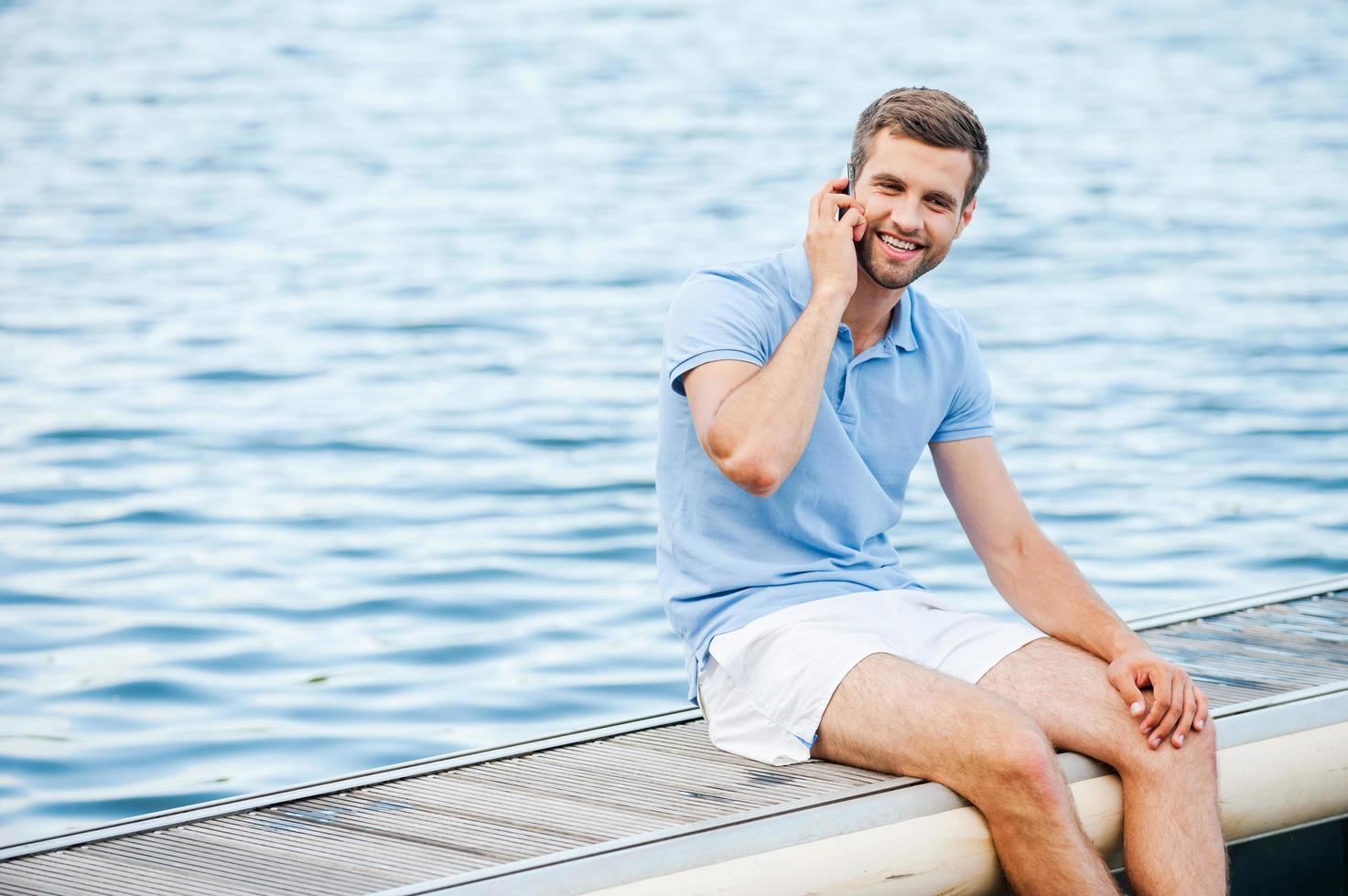 vistas ansluten överallt. stilig ung man i polo skjorta talande på de mobil telefon och leende medan Sammanträde på kajkant foto