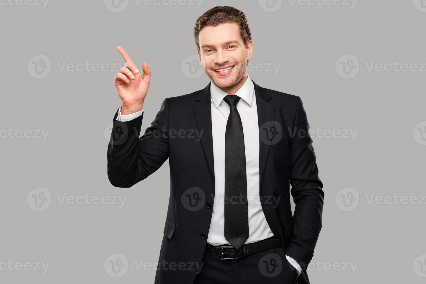 reklam din produkt. självsäker ung man i formell klädsel ser på kamera och leende medan pekande bort och stående mot grå bakgrund foto