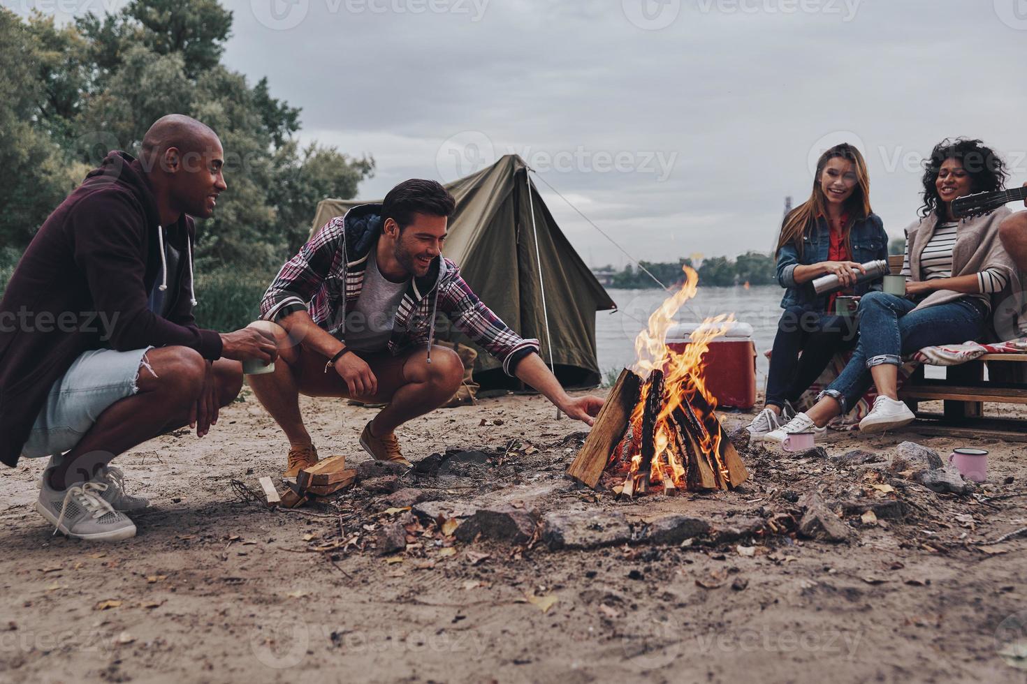 helt enkelt avkopplande. grupp av ung människor i tillfällig ha på sig leende medan njuter strand fest nära de lägereld foto