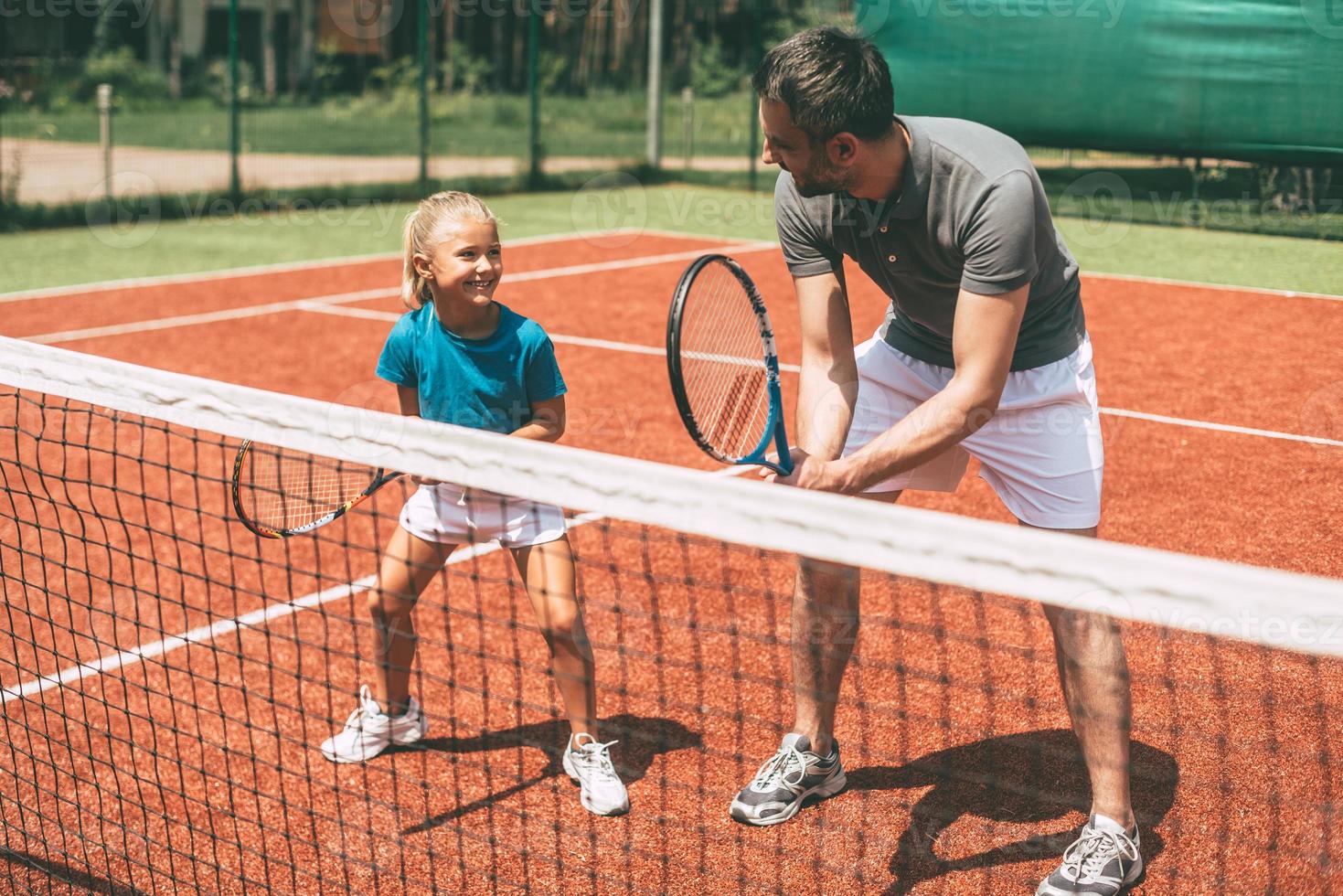 tennis Träning. glad far i sporter Kläder undervisning hans dotter till spela tennis medan både stående på tennis domstol foto