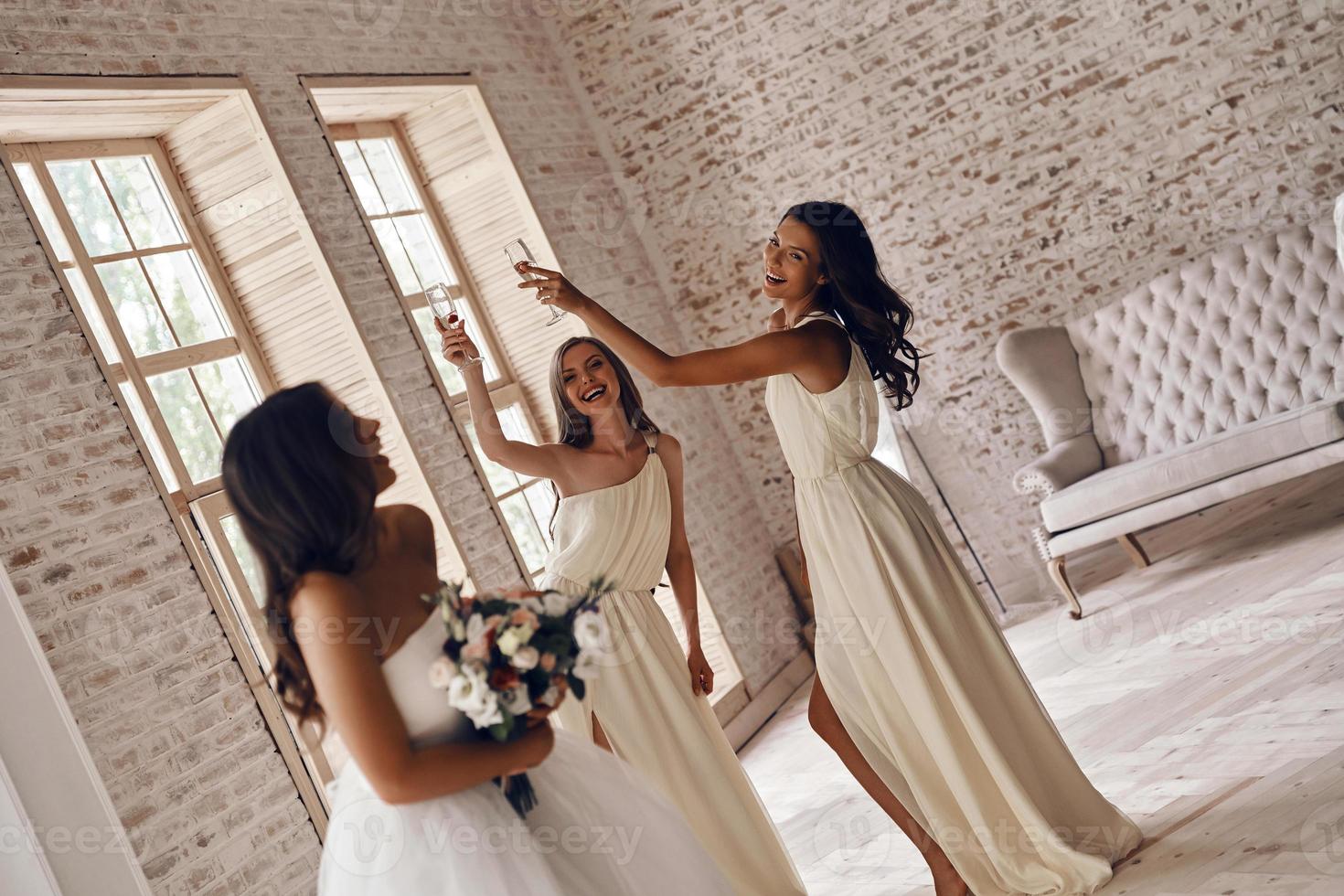 attraktiv ung kvinna innehav en bröllop bukett och leende medan henne flick dricka champagne i de bakgrund foto