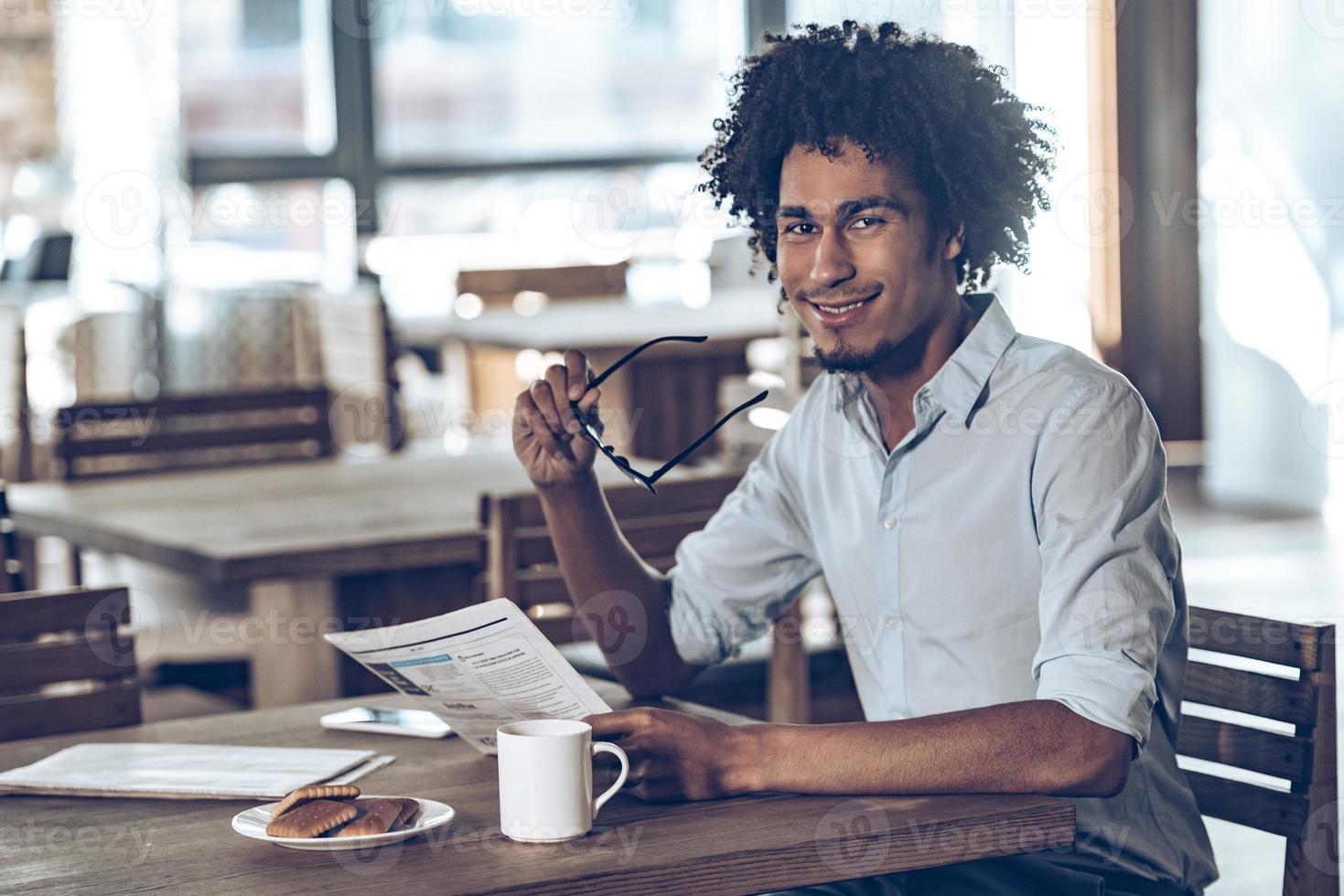 Hej där ung afrikansk man innehav tidning och ser på kamera med leende medan Sammanträde i Kafé foto