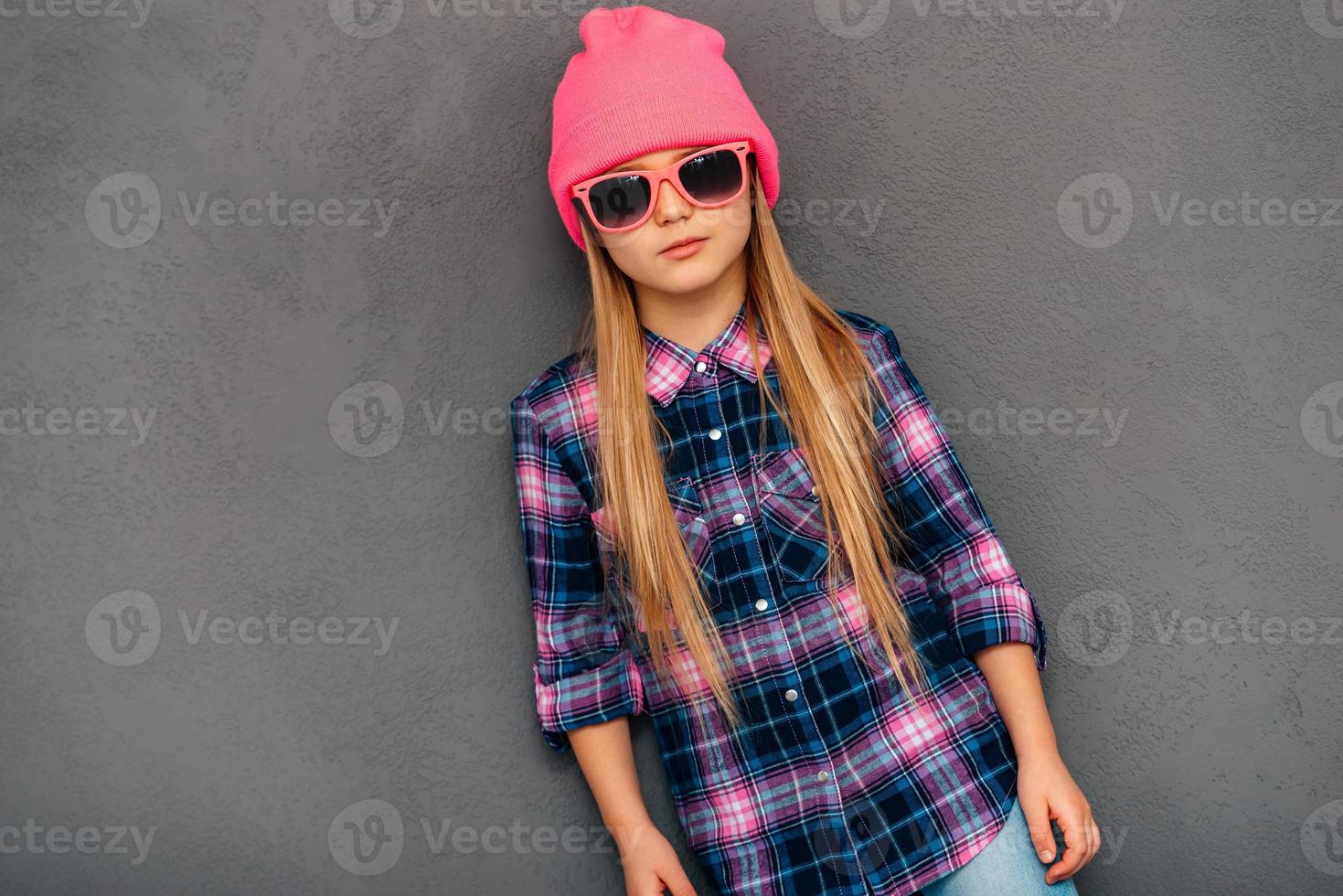självsäker liten Fröken. vacker liten flicka i solglasögon och mössa hatt ser på kamera medan stående mot grå bakgrund foto