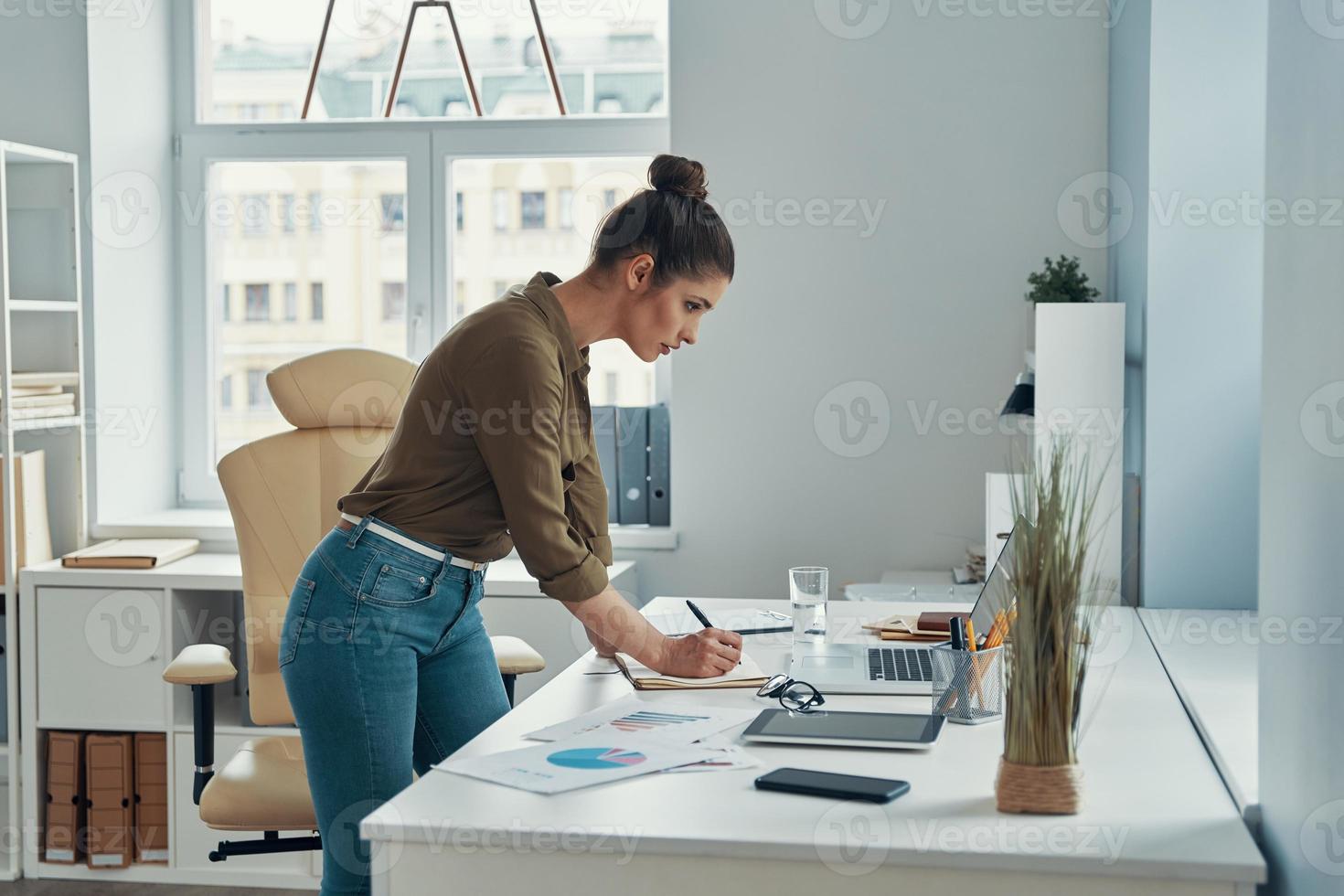 skön ung kvinna i smart tillfällig ha på sig skrivning något ner medan stående i de kontor foto
