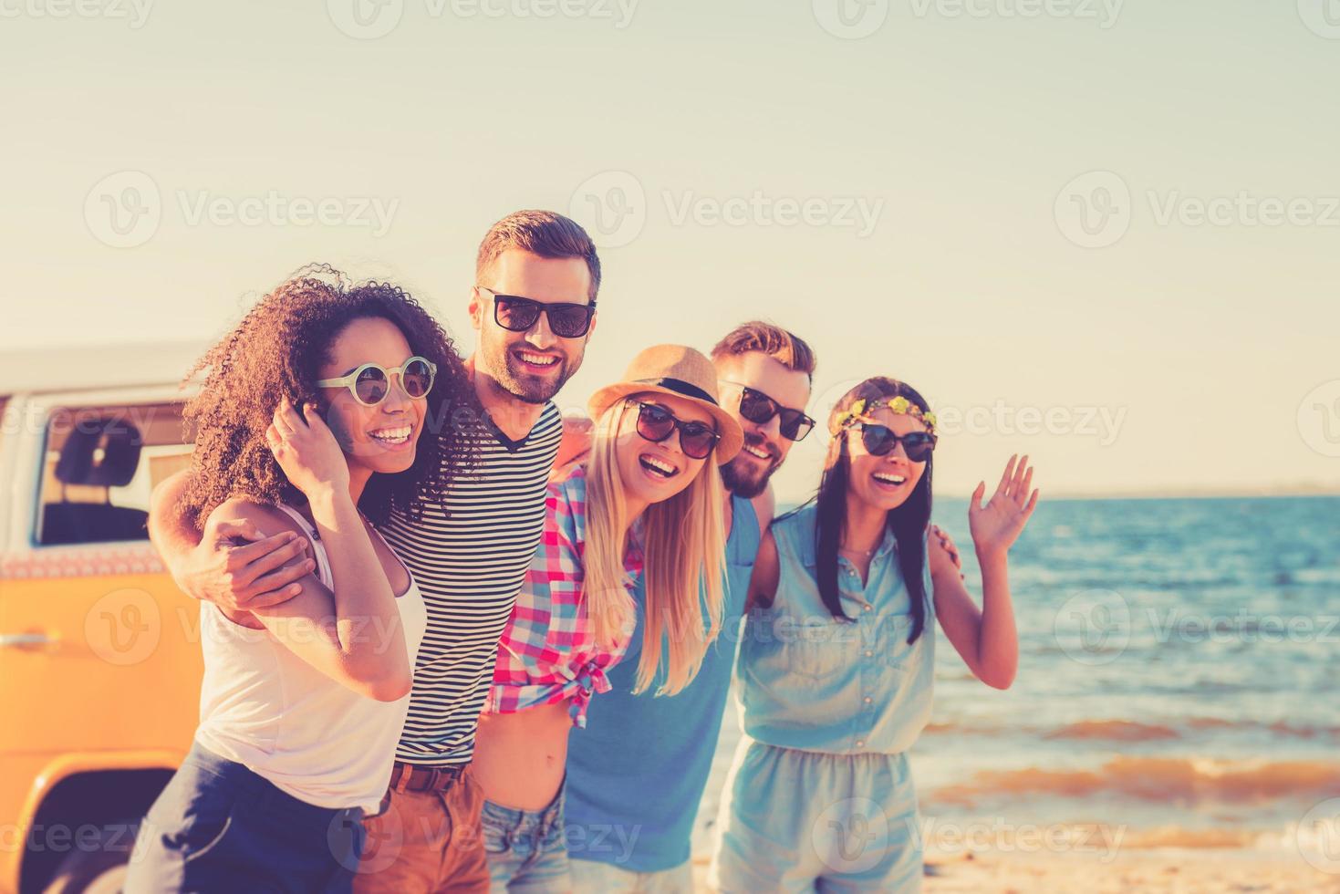 njuter frihet. grupp av glad ung människor fattande och ser på kamera medan gående längs de strand foto