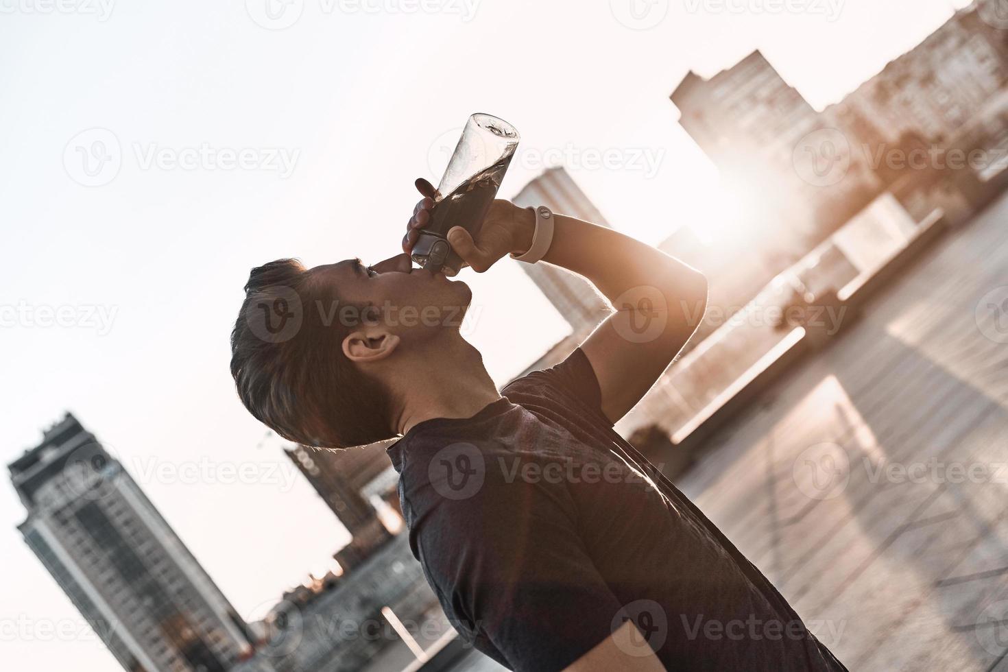 återfuktande. ung man i sporter Kläder dricka vatten medan stående utanför foto