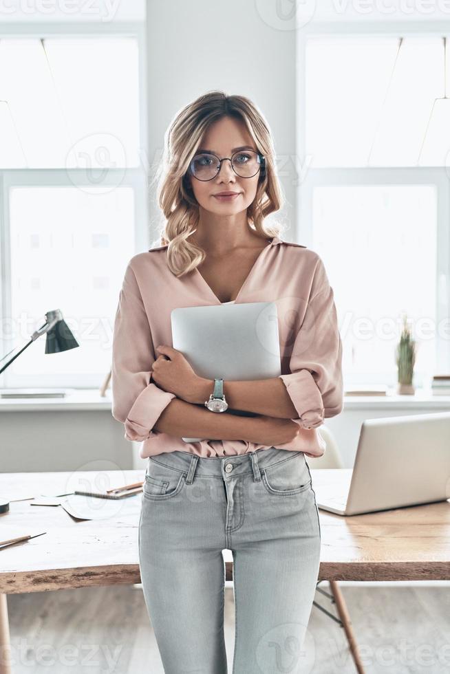 självsäker och framgångsrik. ung attraktiv kvinna ser på kamera och innehav digital läsplatta medan stående i modern kontor foto