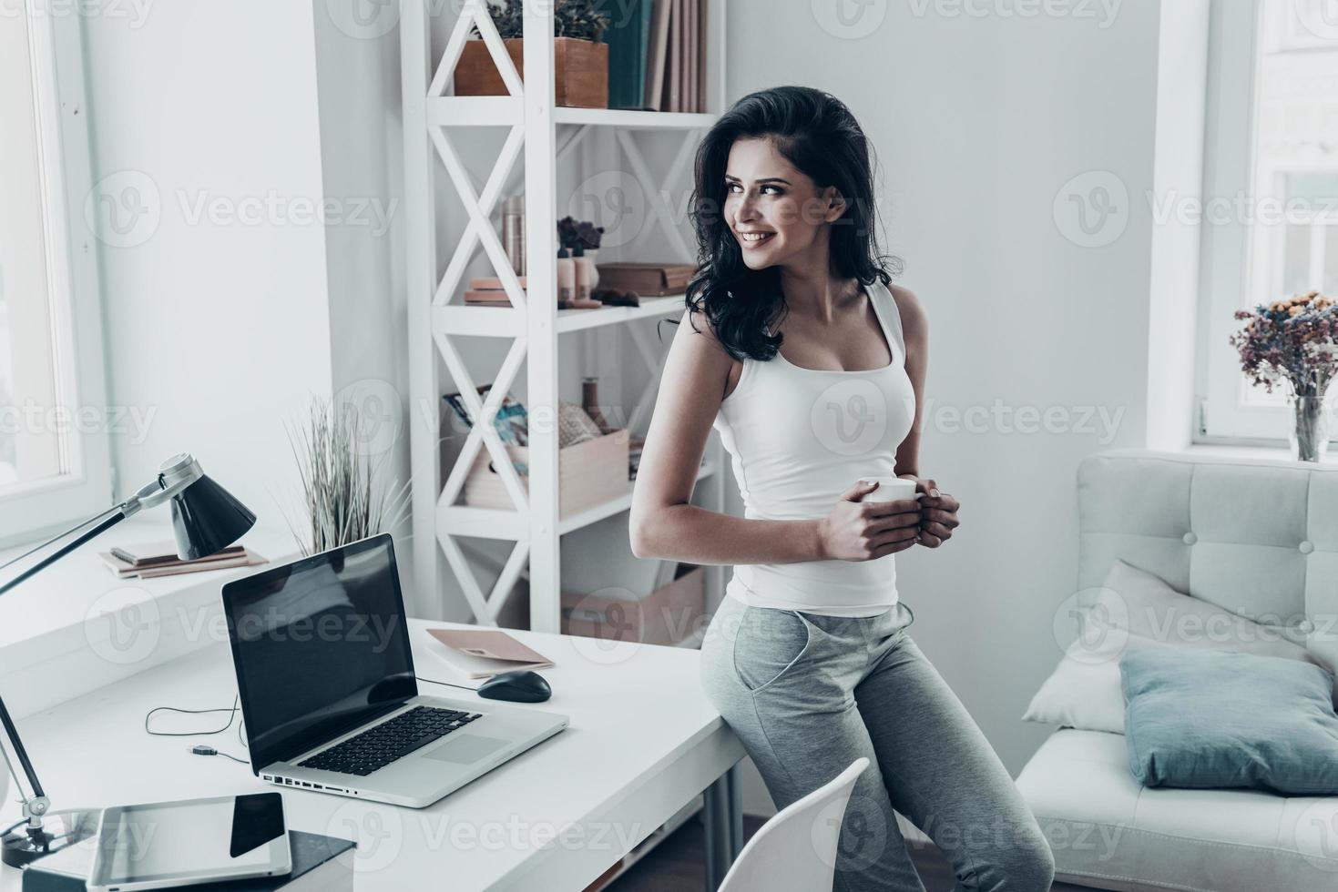 i Bra humör. skön ung kvinna ser utanför och innehav en kopp medan lutande på de skrivbord på Hem foto