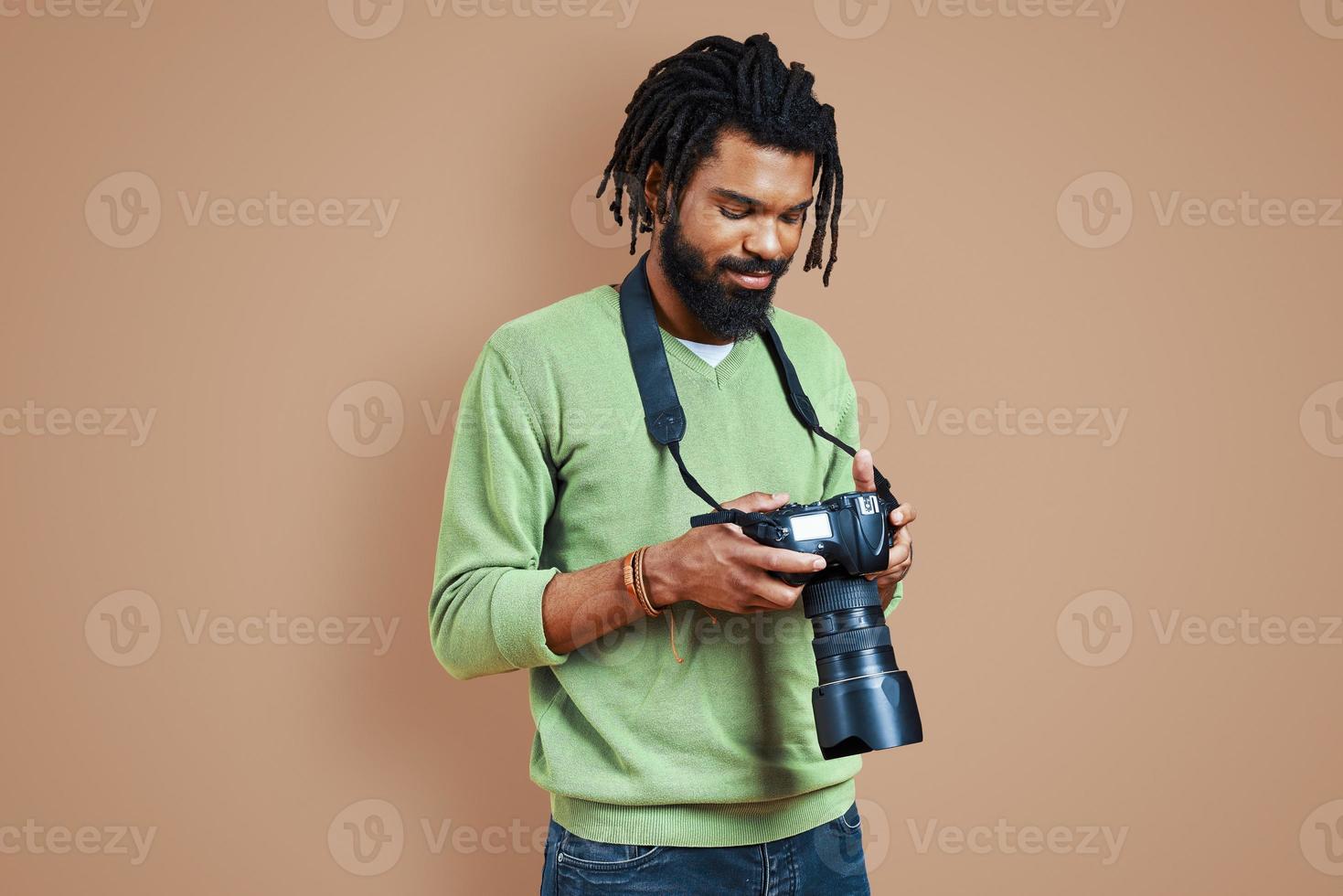 stilig ung afrikansk fotograf i tillfällig Kläder använder sig av digital kamera och leende medan stående mot brun bakgrund foto