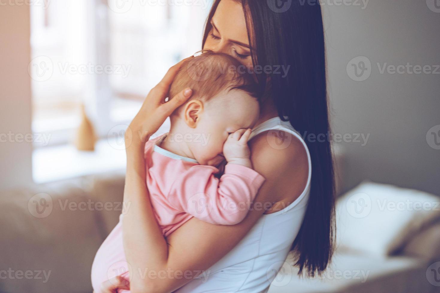 sömnig bebis. sida se av skön ung kvinna innehav sömnig bebis flicka i henne vapen och bindning med henne medan stående på Hem foto