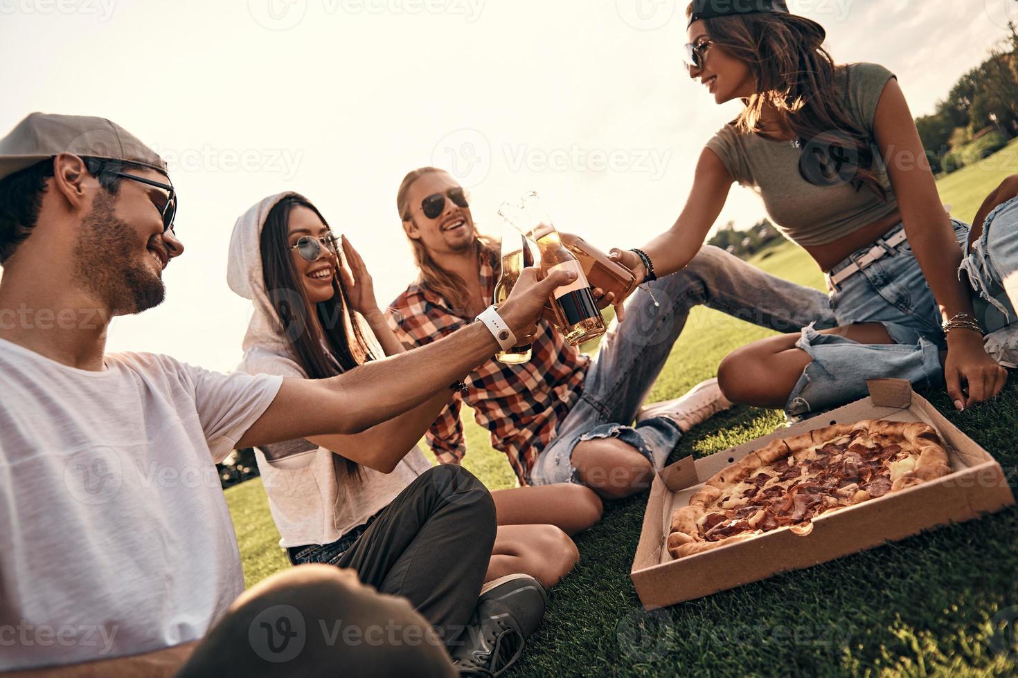 ung och sorglös. grupp av leende människor i tillfällig ha på sig toasting med öl flaskor medan njuter picknick utomhus foto