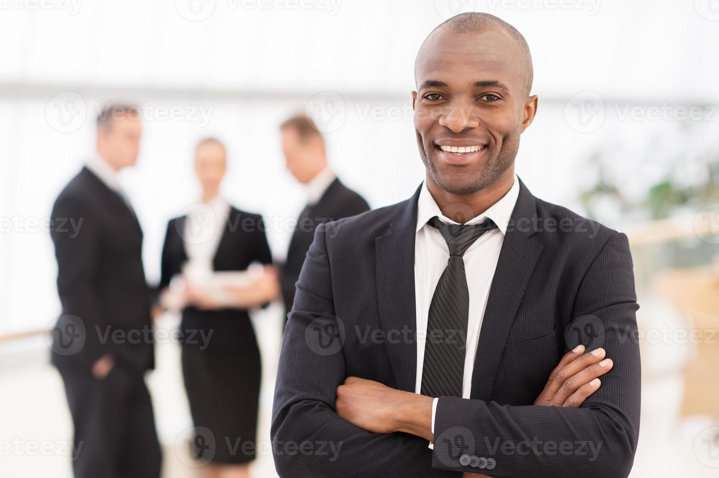 självsäker affärsman. glad ung afrikansk man i formell klädsel förvaring vapen korsade och leende medan hans kollegor stående på bakgrund foto