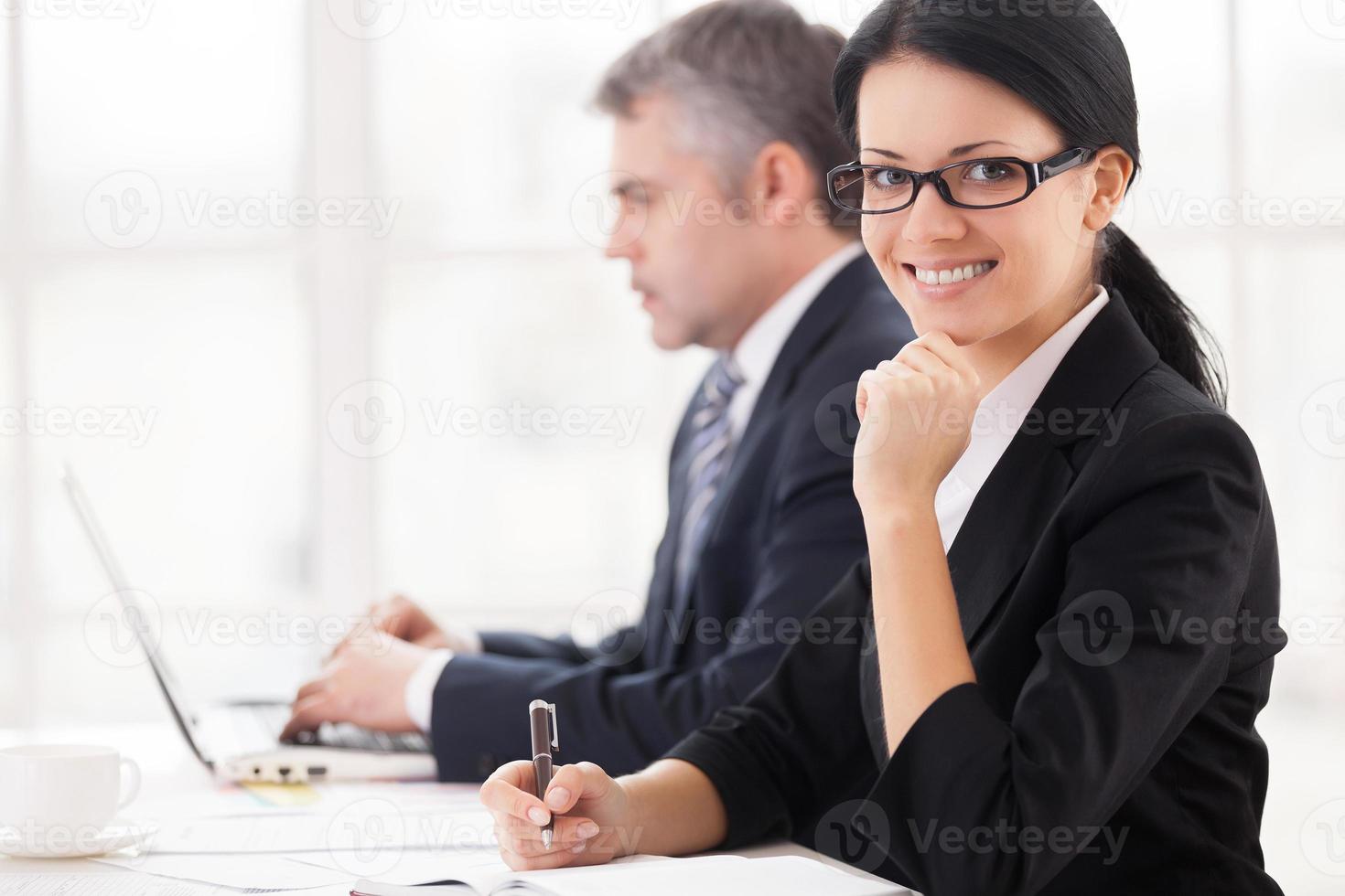 företag människor på arbete. glad ung affärskvinna Sammanträde på de tabell och leende på kamera medan mogna man arbetssätt på bärbar dator på bakgrund foto