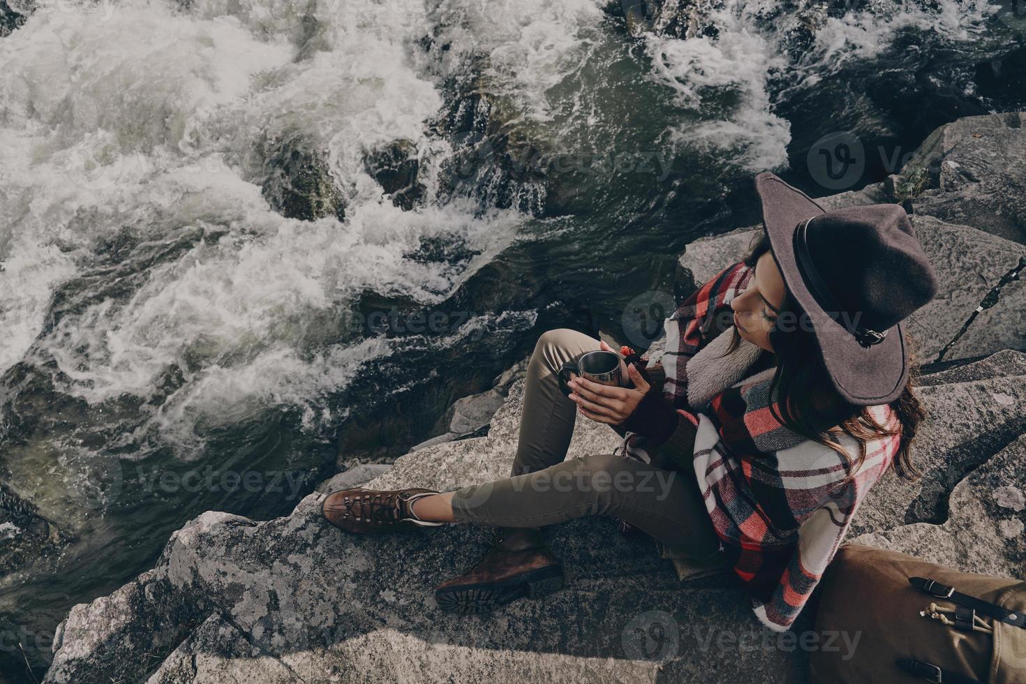 ganska begrundande. topp se av skön ung kvinna täckt med filt innehav en råna medan Sammanträde nära de flod i bergen foto