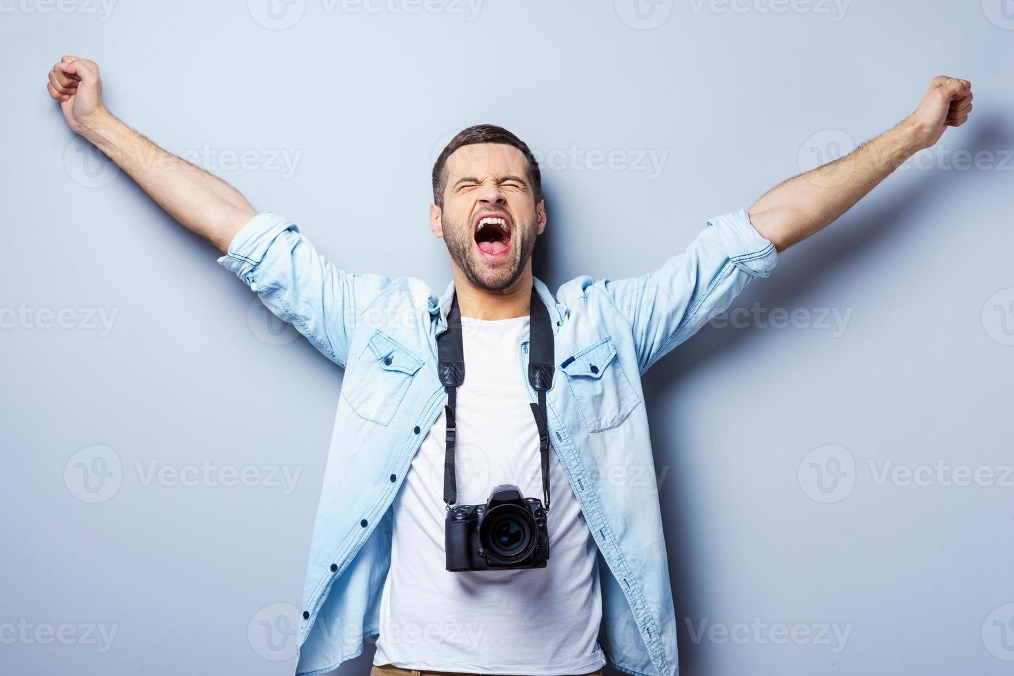 framgångsrik fotograf. Lycklig ung man med digital kamera förvaring vapen Uppfostrad och ögon stängd medan stående mot grå bakgrund foto