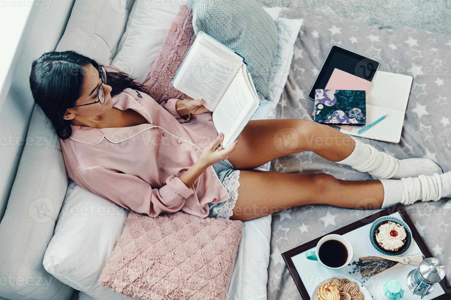 topp se av skön ung kvinna i pyjamas läsning bok medan vilar i säng på Hem foto