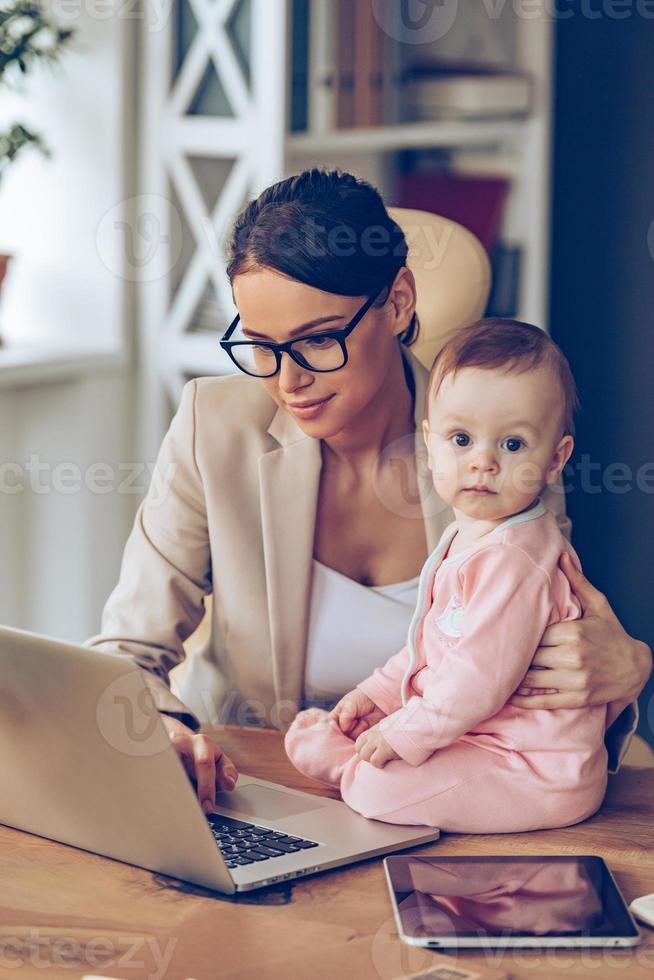 min mamma är en verklig expert- liten bebis flicka ser på kamera medan Sammanträde på kontor skrivbord med henne mor i kontor foto