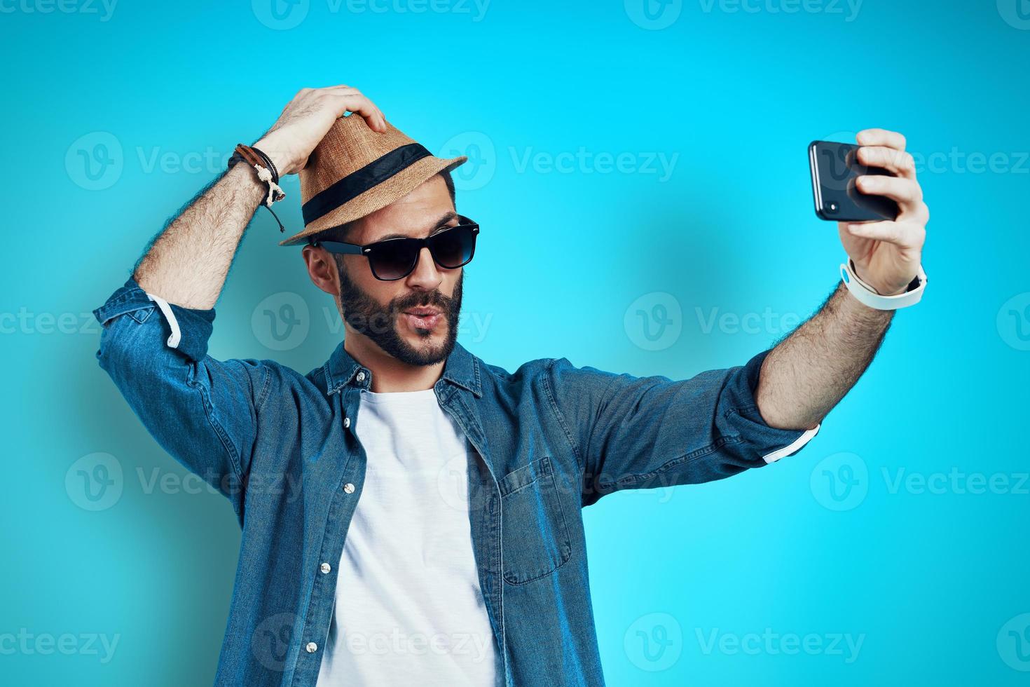 Bra ser ung man leende och tar selfie använder sig av telefon medan stående mot blå bakgrund foto