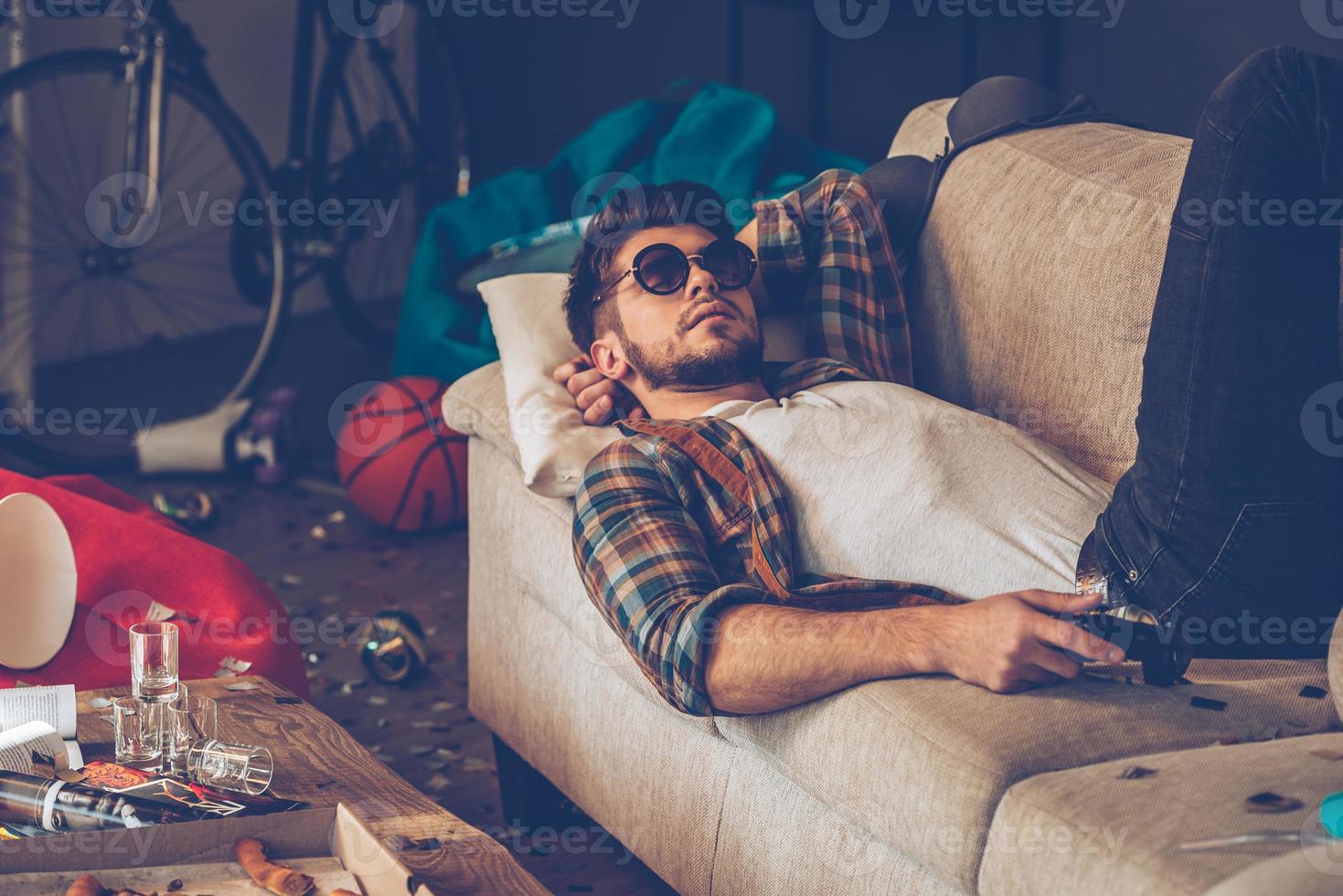 avkopplande efter fest. ung stilig man i solglasögon liggande ner på soffa med joystick i hans hand i rörig rum efter fest foto