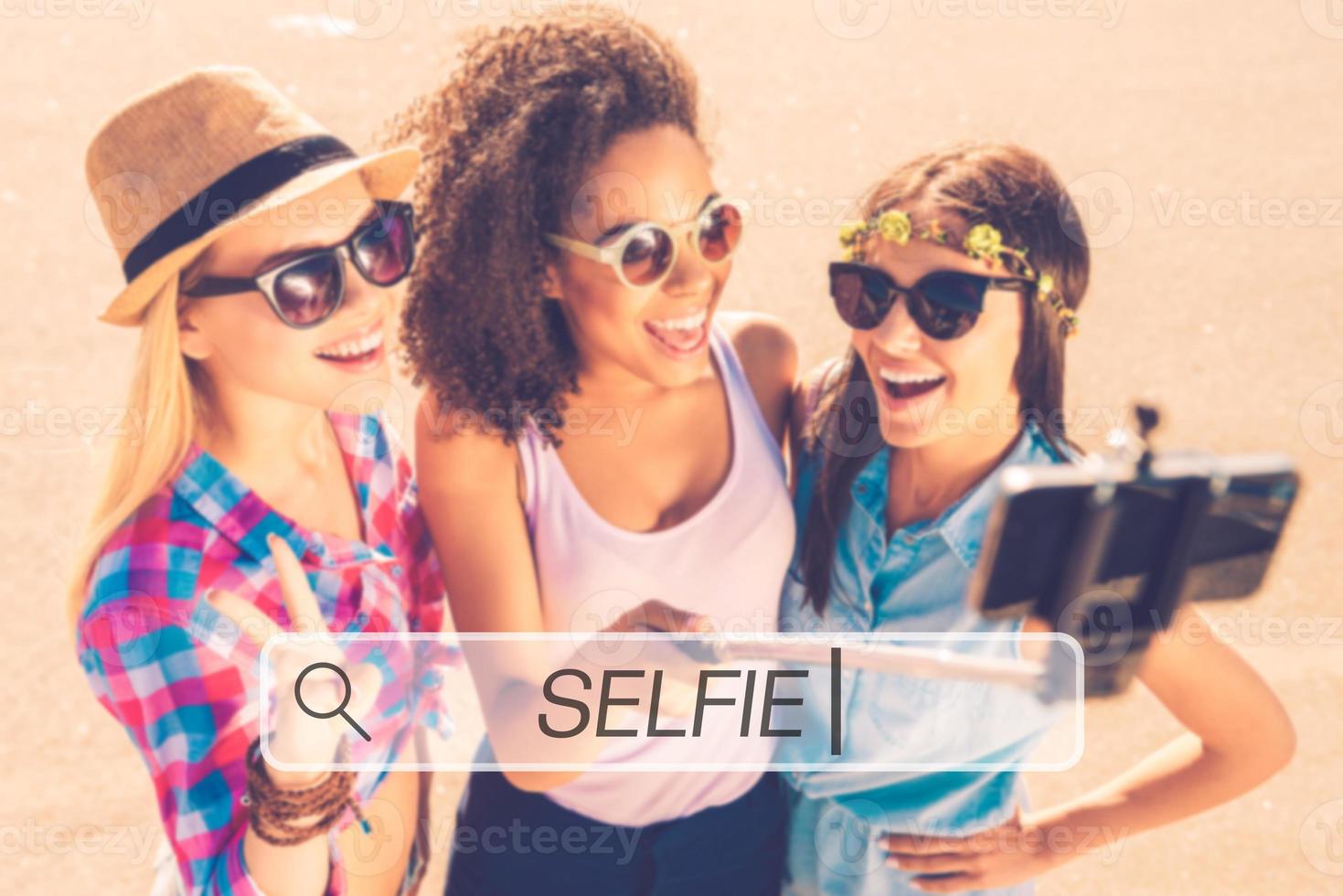 selfie tid topp se av tre ung Lycklig kvinnor framställning selfie förbi deras smart telefon medan stående utomhus tillsammans foto