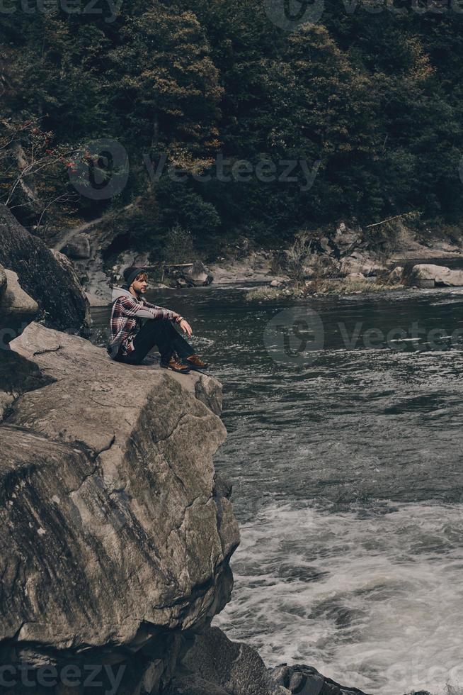 känsla de kraft av natur. stilig ung man njuter de se medan Sammanträde på de sten nära de flod foto