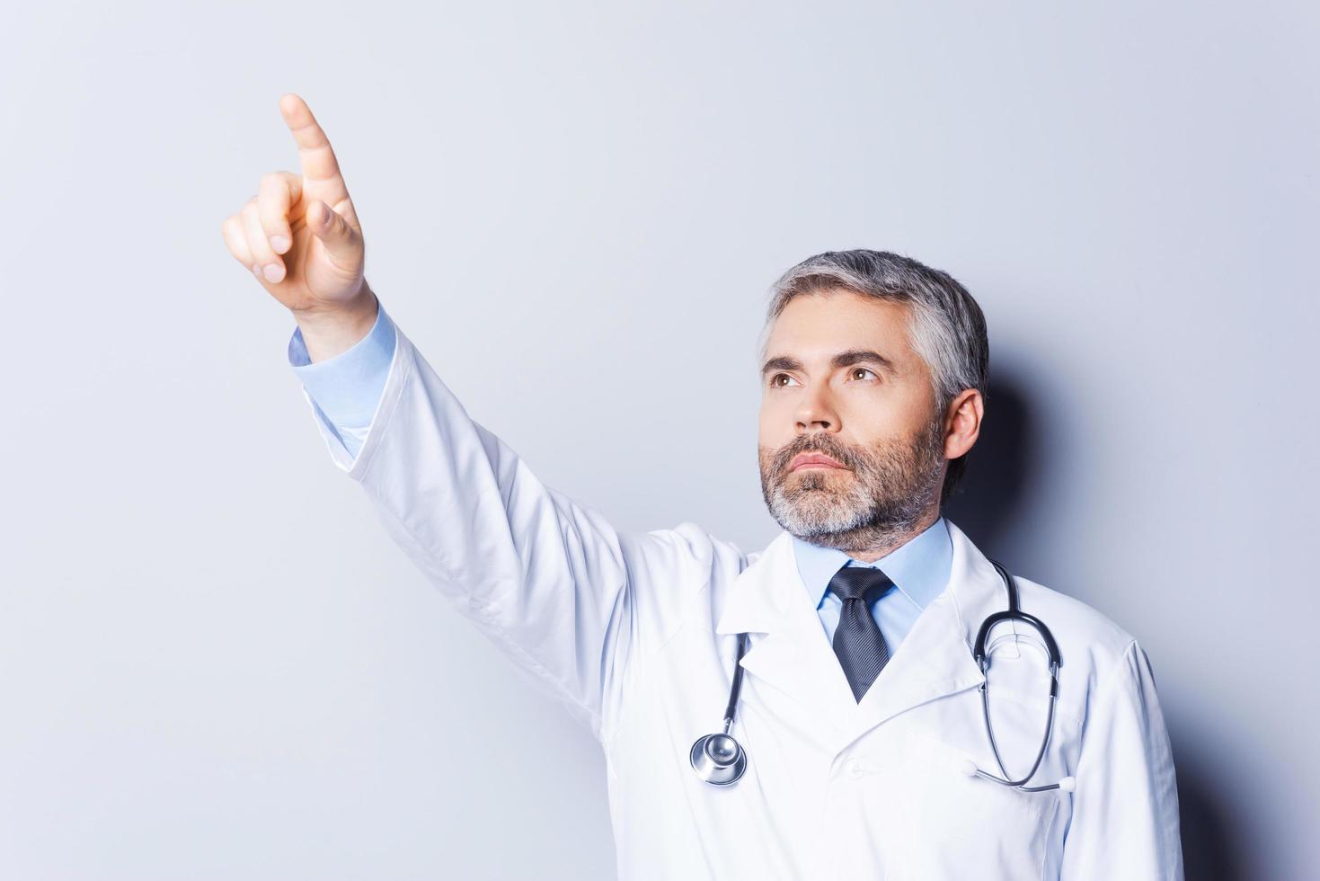 läkare pekande bort. självsäker mogna grå hår läkare pekande bort medan stående mot grå bakgrund foto