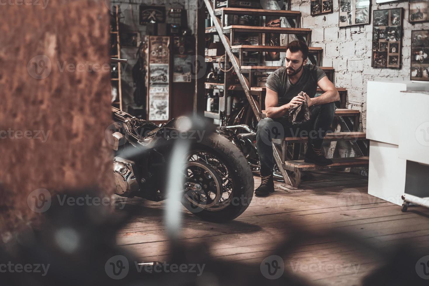 han behov en liten ha sönder. självsäker ung man innehav trasa och ser på motorcykel medan Sammanträde nära den i reparera affär foto