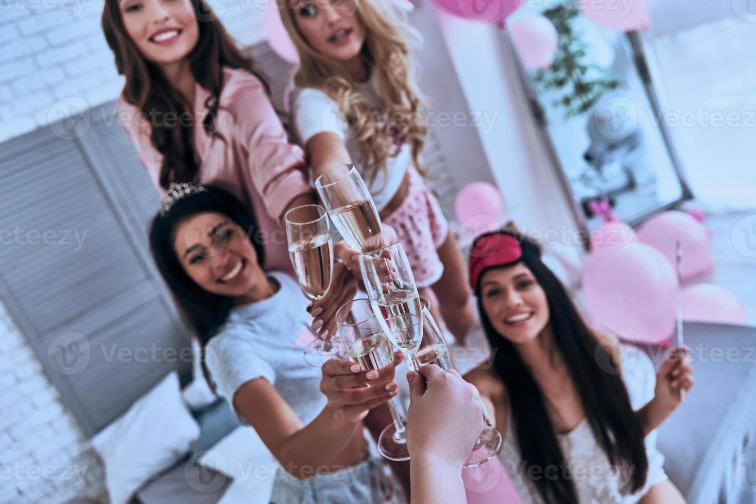 Skål till oss fyra lekfull ung leende kvinnor i pyjamas toasting varje Övrig medan har en slummer fest i de sovrum med ballonger Allt över de plats foto