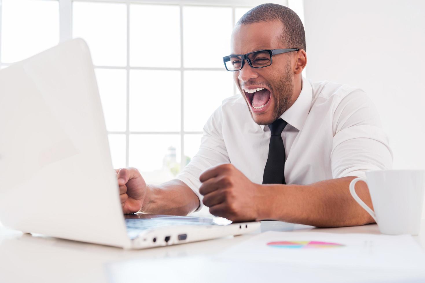 för stressig arbete. rasande ung afrikansk man i skjorta och slips skrikande medan ser på bärbar dator medan Sammanträde på hans arbetssätt plats foto