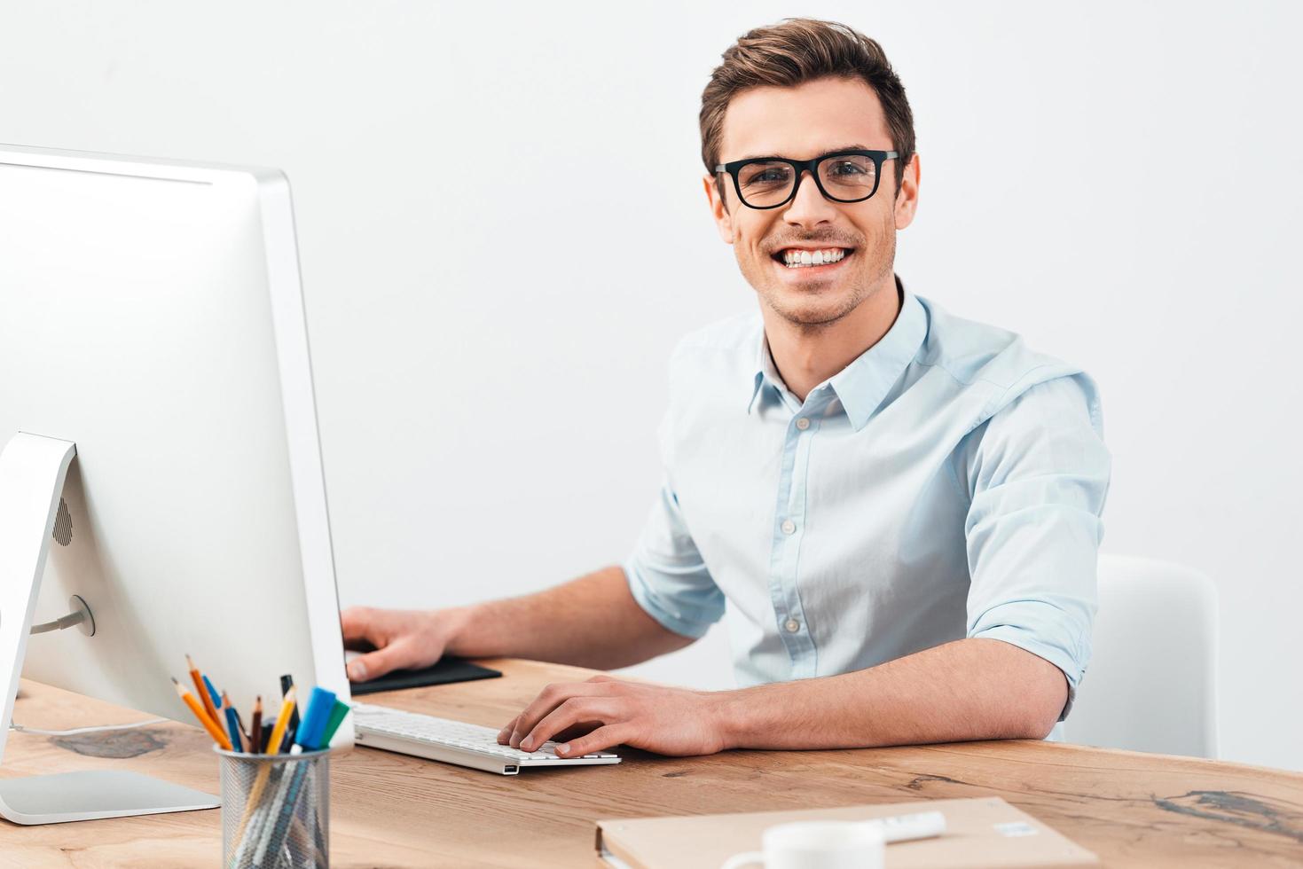 bäst chef. glad ung stilig man i glasögon arbetssätt på dator och ser på kamera med leende medan Sammanträde på hans arbetssätt plats foto