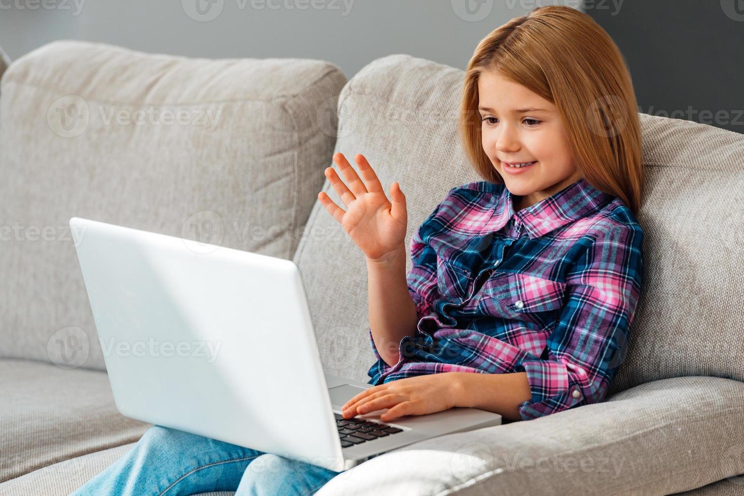 Hej mormor skön liten flicka använder sig av henne bärbar dator och vinka med henne hand medan Sammanträde på de soffa på Hem foto