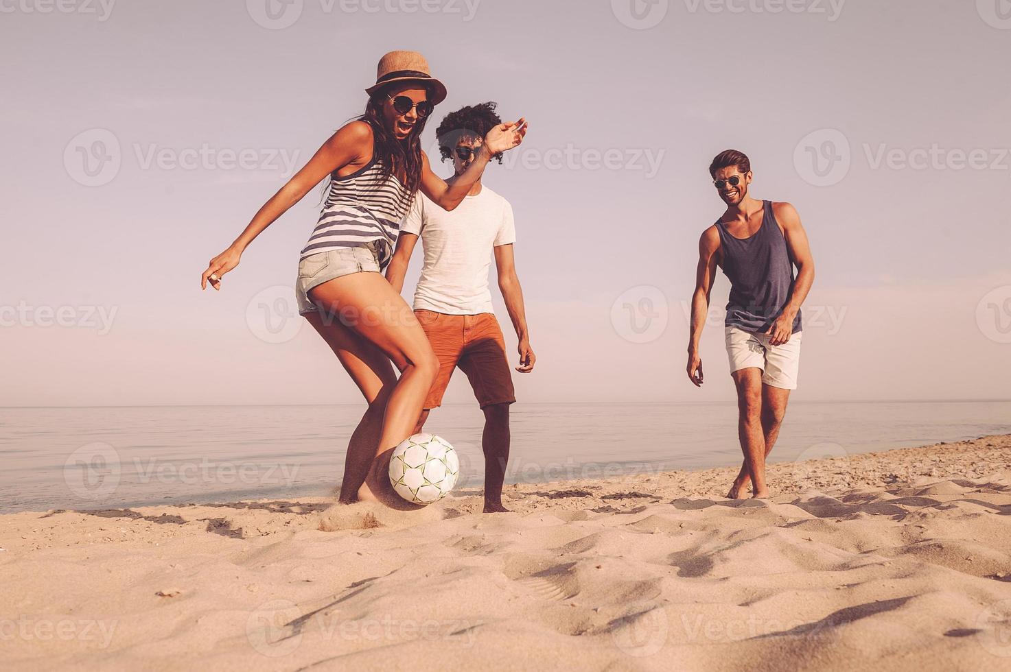 roligt tid med vänner. tre glad ung människor spelar med fotboll boll på de strand foto