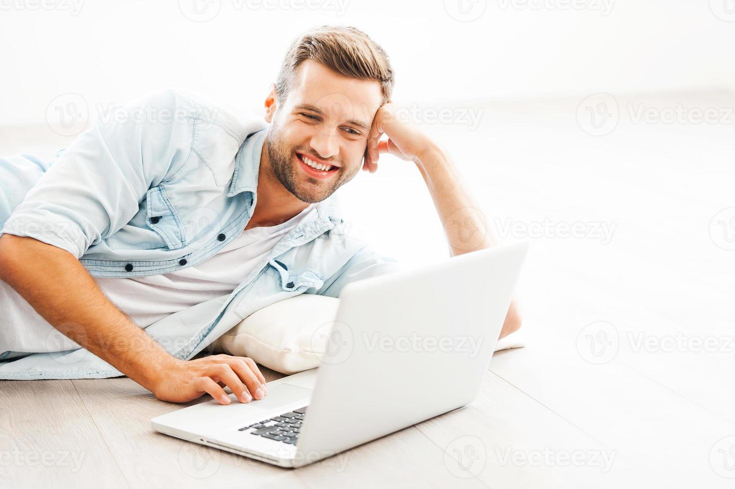 avkopplande på Hem. glad ung man arbetssätt på bärbar dator och leende medan liggande på de golv på hans lägenhet foto