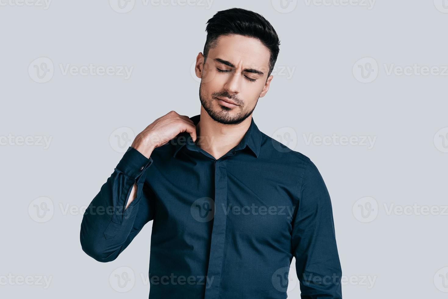 känsla trött. utmattad ung man dragande krage av hans skjorta med stängd ögon medan stående mot grå bakgrund foto