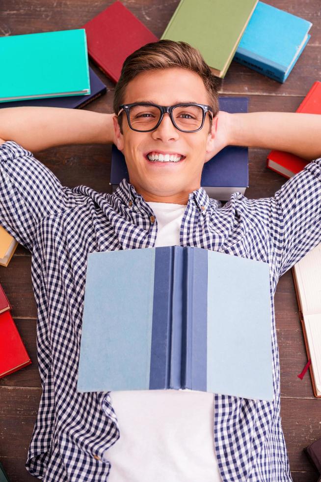 Lycklig bokmal. topp se av Lycklig ung man innehav händer Bakom huvud och leende medan liggande på de hårt träslag golv med färgrik böcker om Allt runt om honom foto
